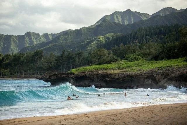 Beach scene with people swimming, a rocky coastline, lush green hills, and mountains in the background under cloudy skies.