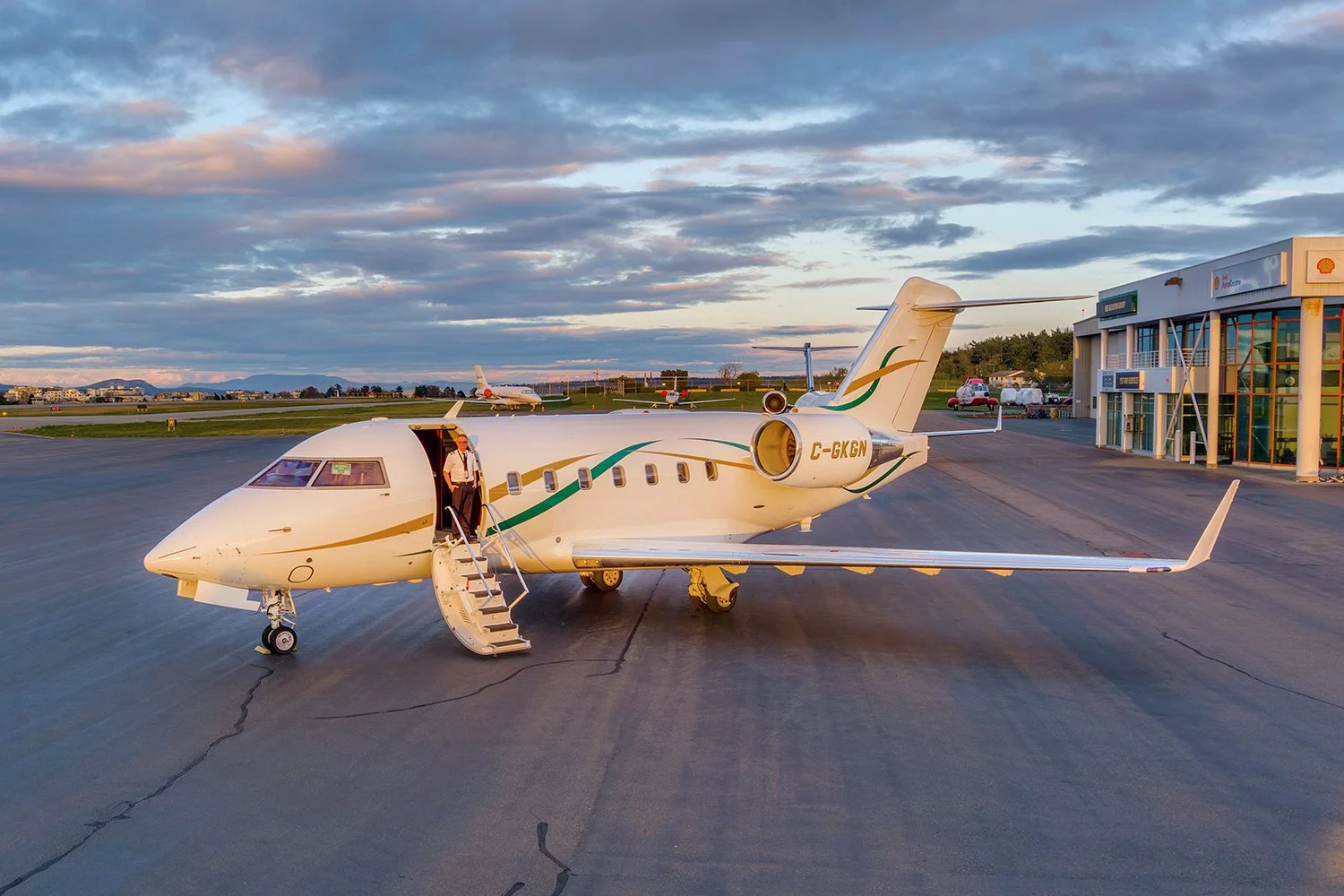 Private jet on tarmac with crew member descending stairs, airport building to the right, other aircraft in distance, cloudy sky, late afternoon or early evening light.