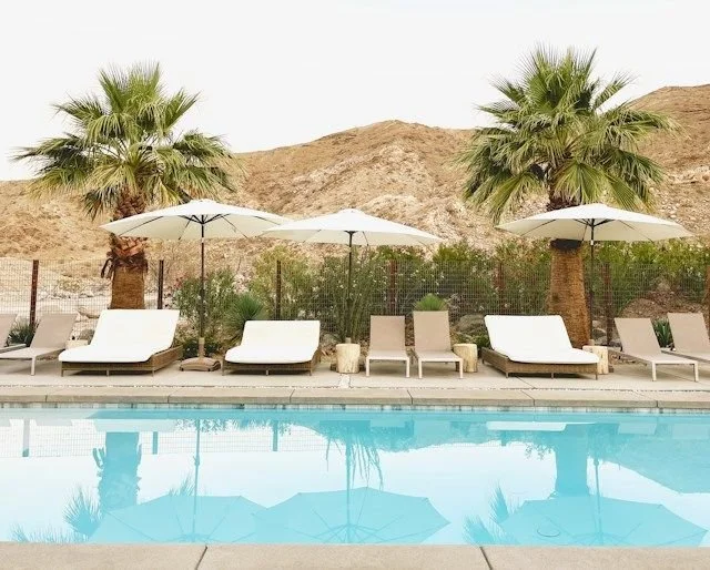Swimming pool with lounge chairs and umbrellas, palm trees, and desert hills in the background.