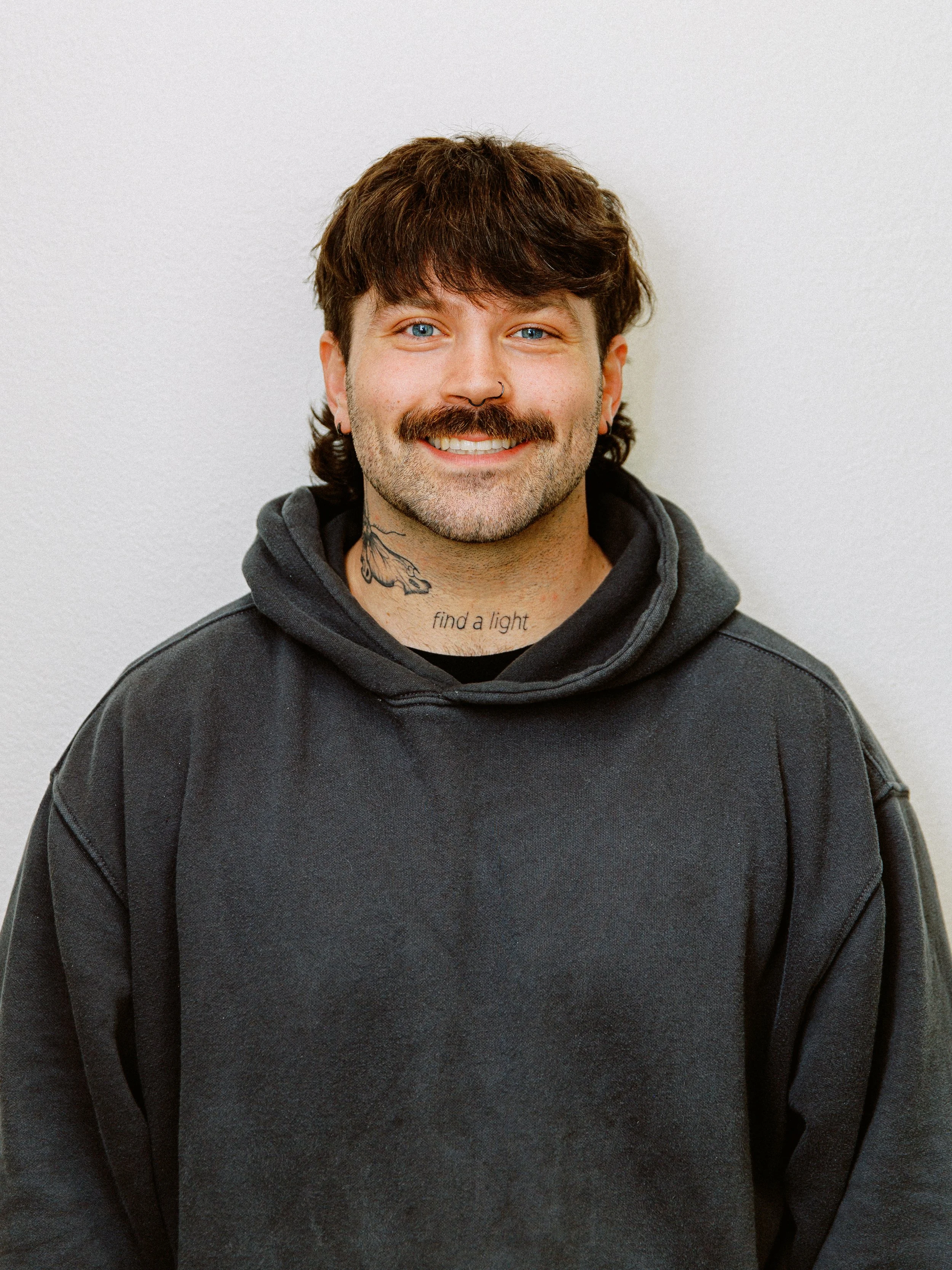 Portrait of a young man with wavy brown hair, a beard, and a big smile, wearing a black button-up shirt against a plain light gray background.