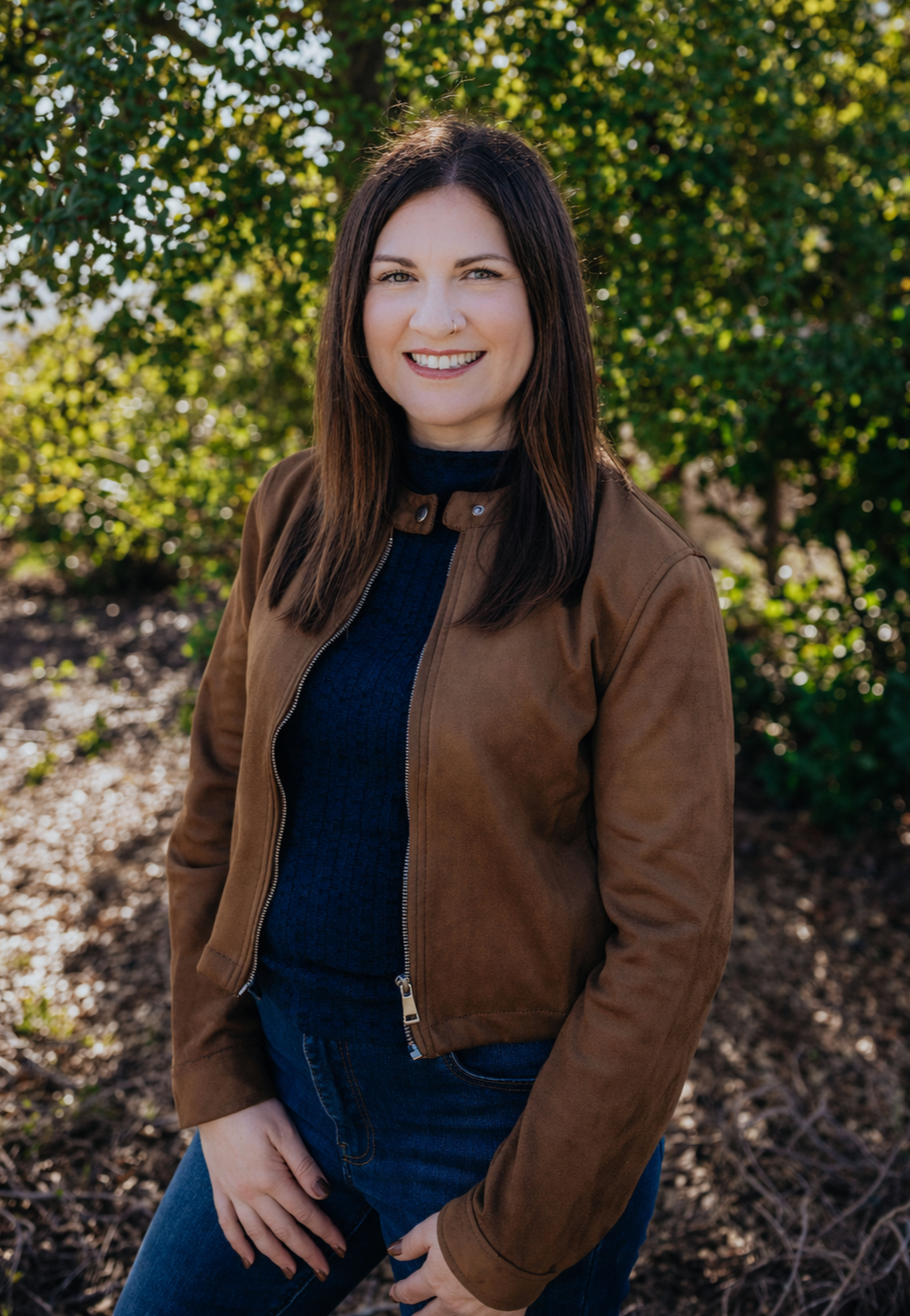 A woman with dark hair, wearing a brown jacket and blue sweater, smiling outdoors with green trees in the background.