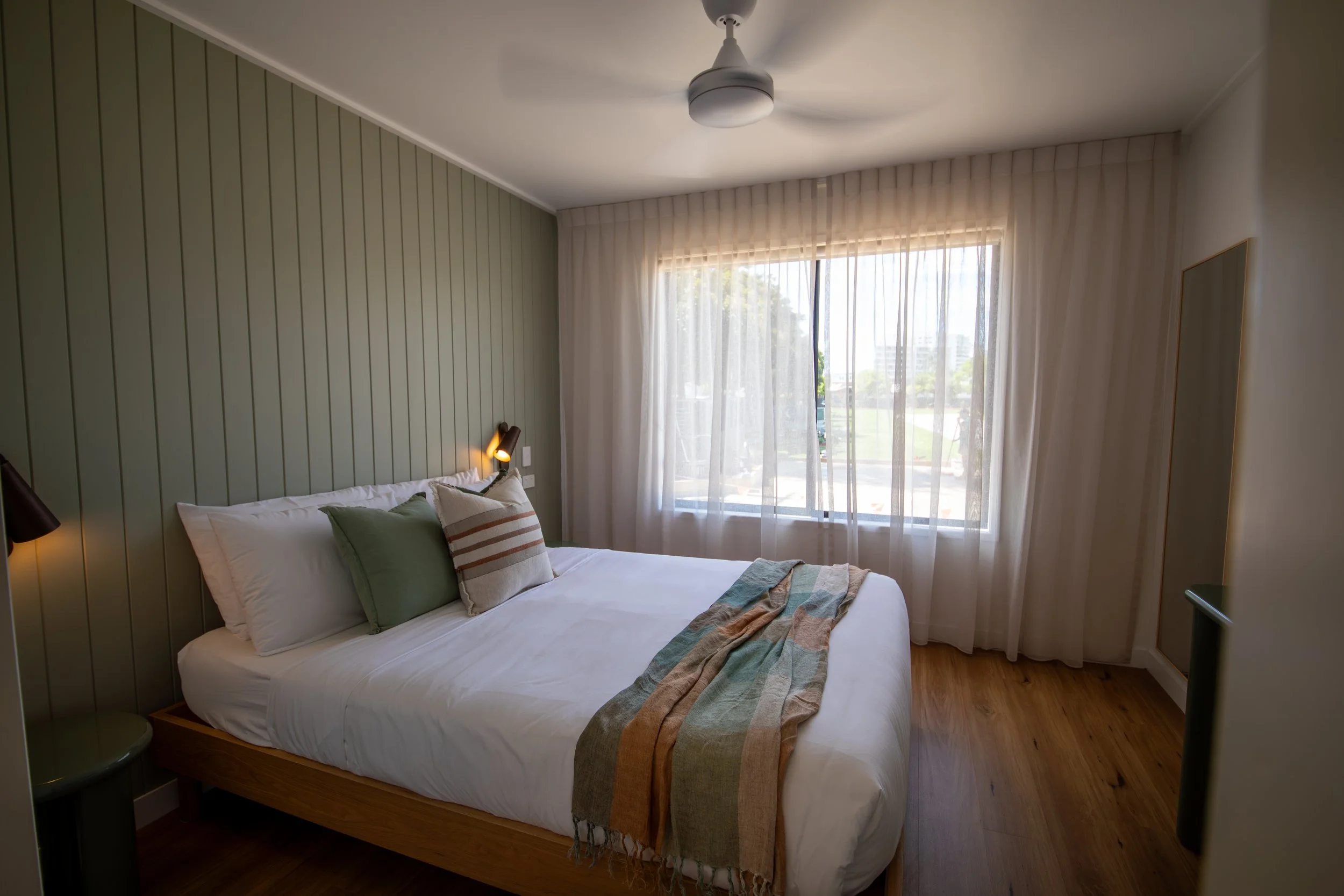 Interior of a small, modern living room and kitchen with white walls, hardwood floors, a window with blinds, a wall-mounted TV, a dining table with four chairs, a gray sofa, a small round coffee table with a plant and remote, a white fridge, and kitchen appliances.