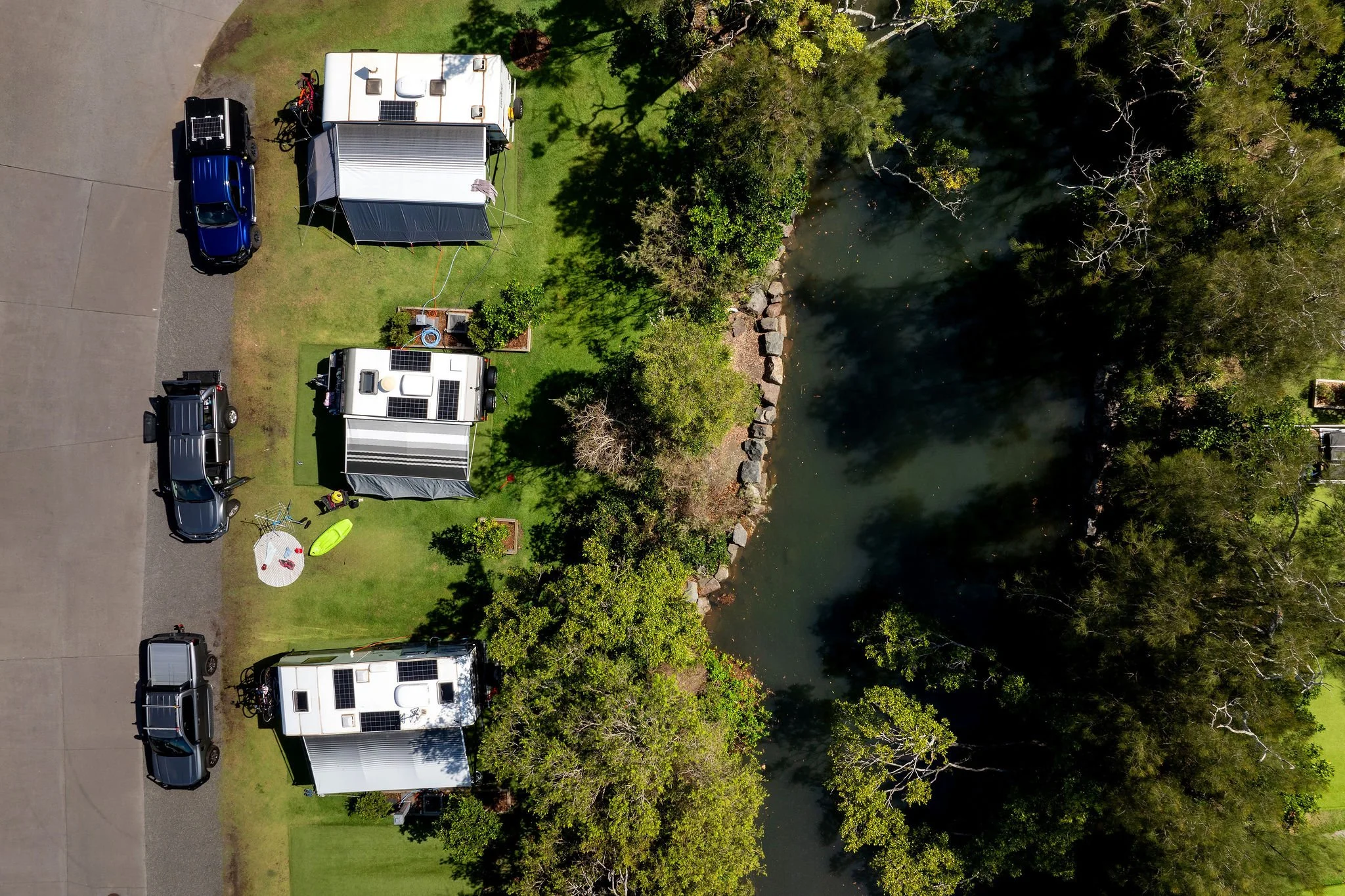 A small square patch of freshly mowed grass surrounded by taller grass, with two mailboxes on a post and a small garden bed with plants nearby, against a backdrop of trees and clear blue sky.