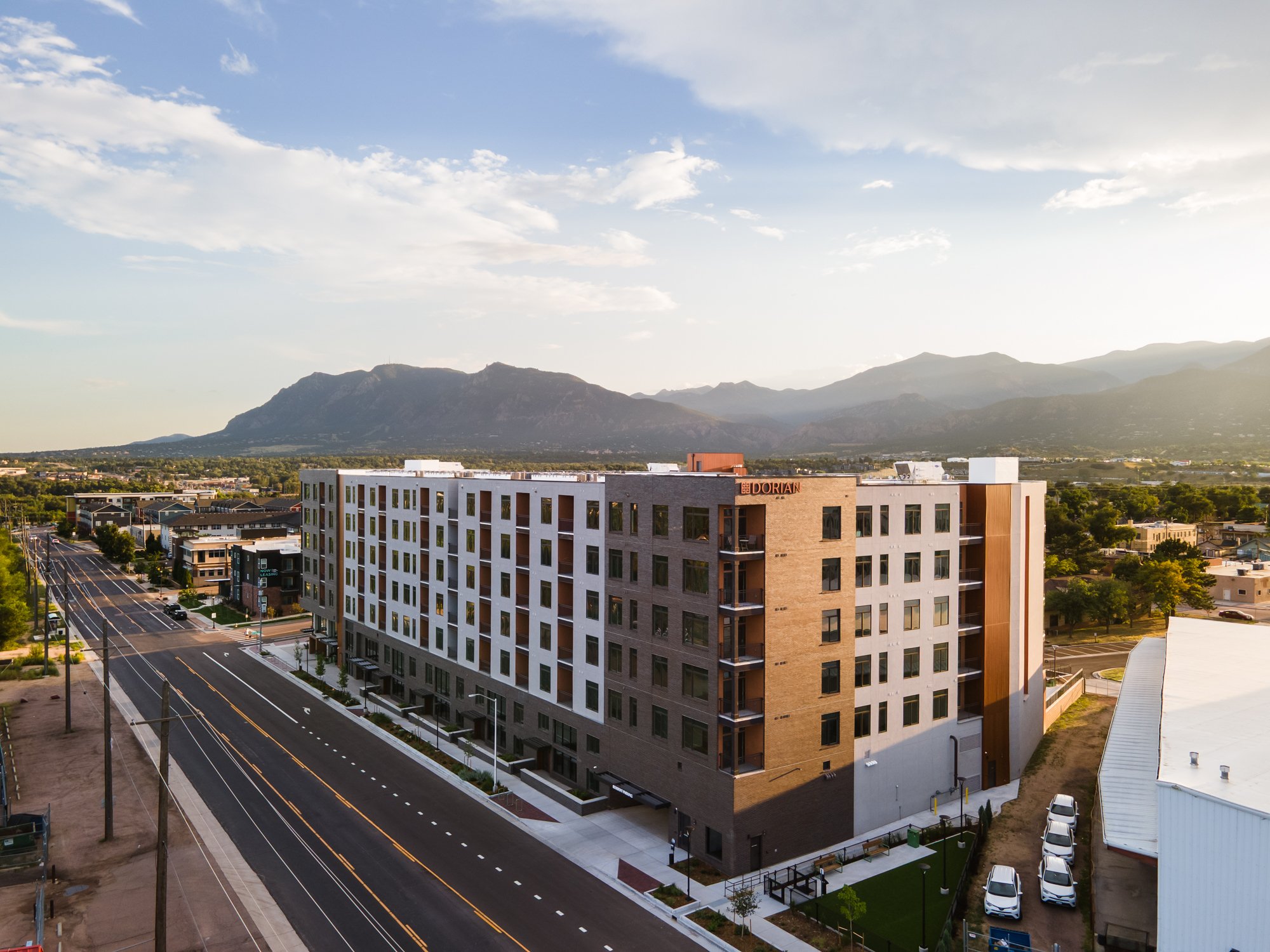 A modern multi-story apartment building with a brick and white exterior, located next to a paved street with cars, set against a backdrop of mountains under a partly cloudy sky.