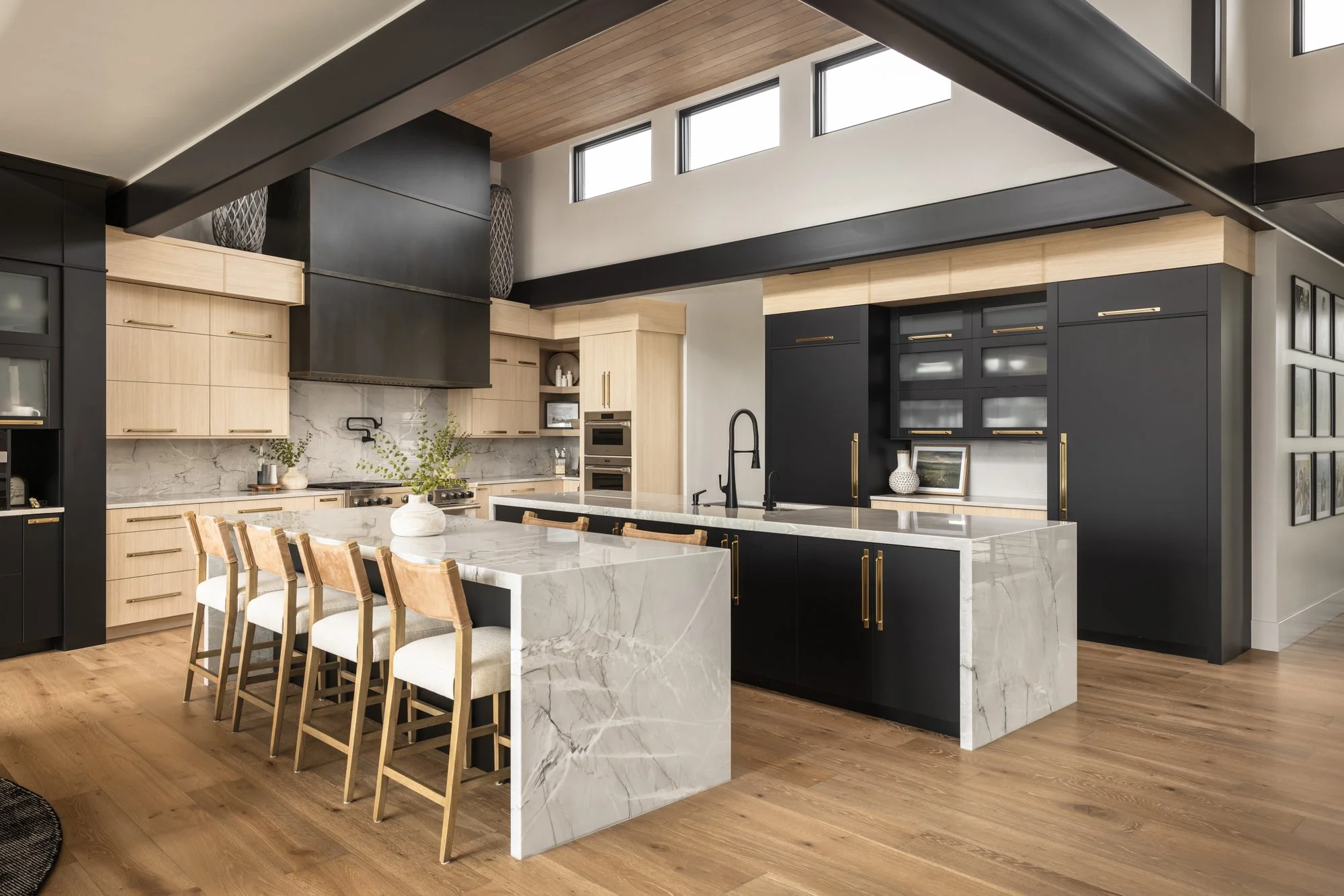 Modern kitchen with black and light wood cabinetry, a large marble island with beige chairs, and wooden flooring.