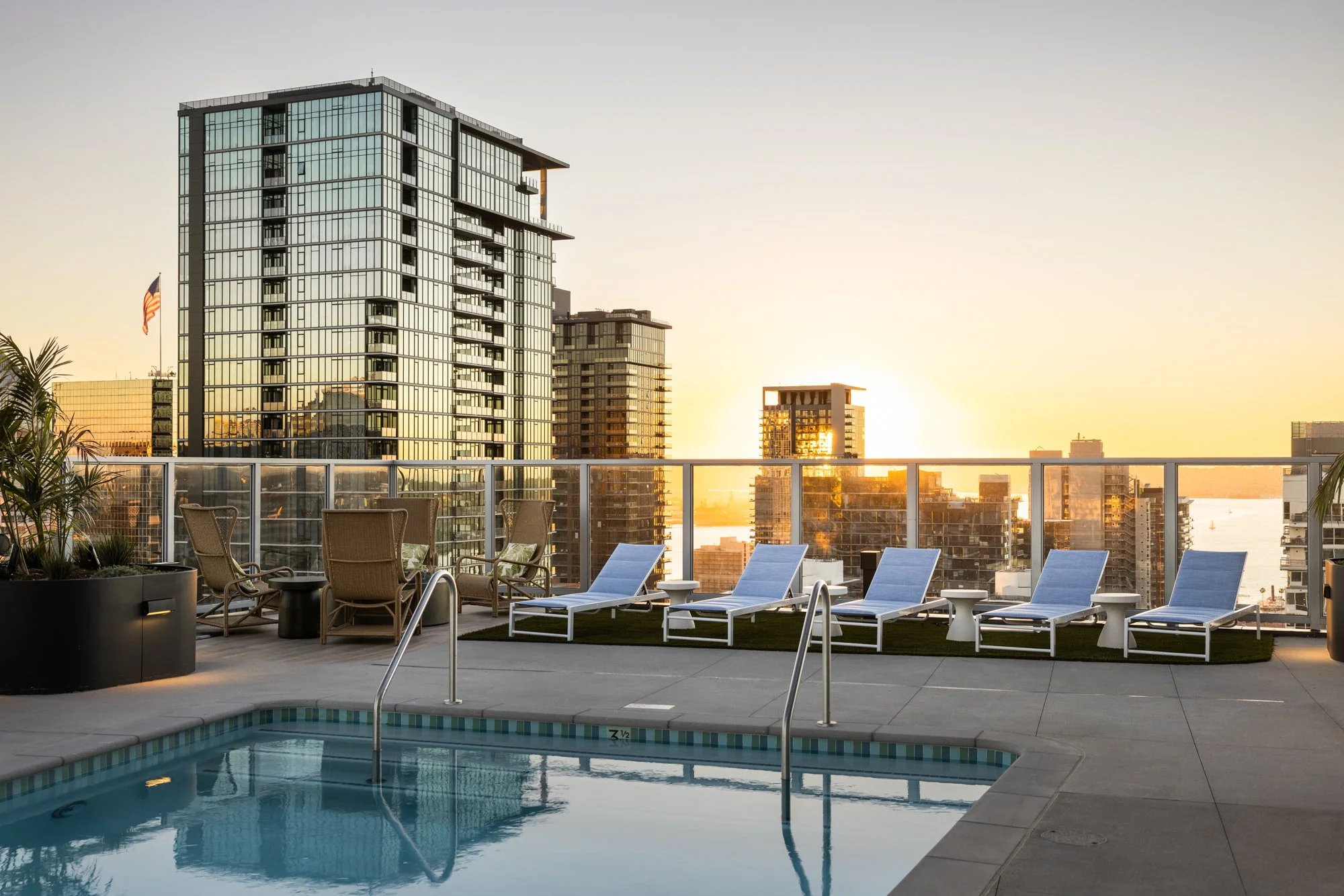 Rooftop swimming pool with lounge chairs, chairs, potted plants, and city skyline at sunset.