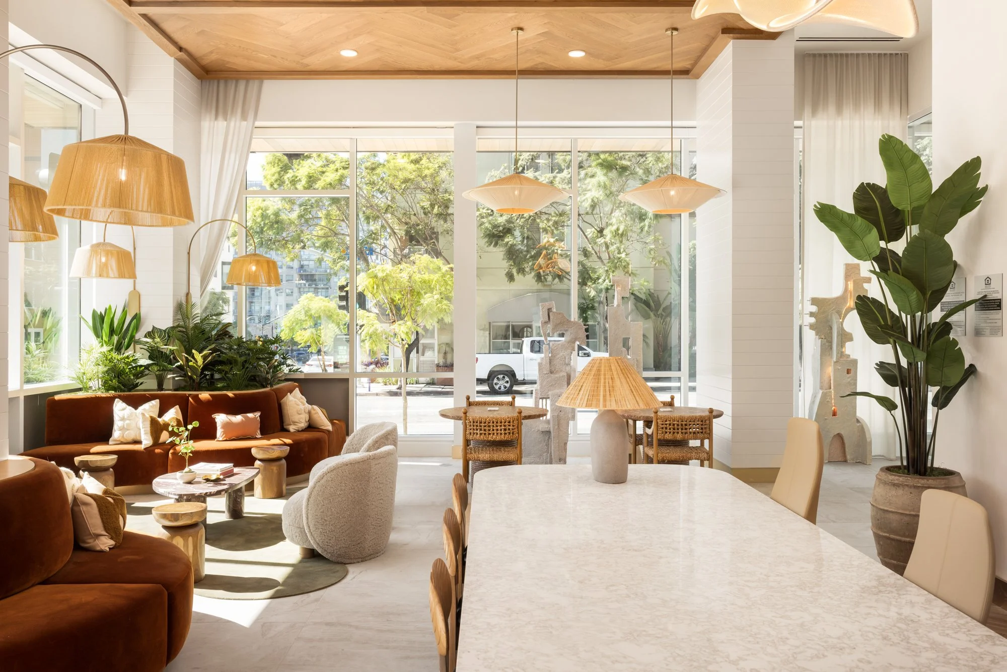 Bright indoor space with large windows, green plants, a brown velvet sofa with pillows, cozy chairs, wooden tables, pendant and table lamps, and a large potted plant in a decorative pot.