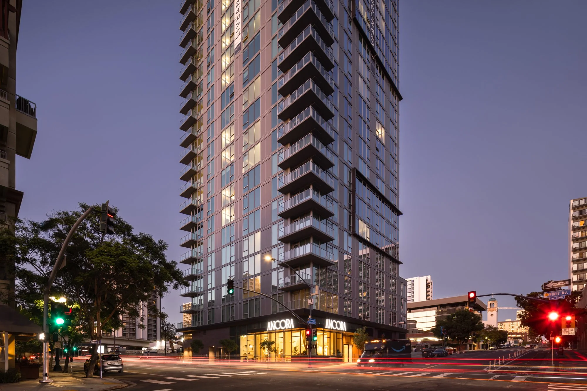 A tall modern high-rise building at dusk with illuminated windows, featuring balconies on each floor. The ground floor has a retail space with the sign "ANCORA". The street below shows traffic lights, cars, and light trails from moving vehicles, with