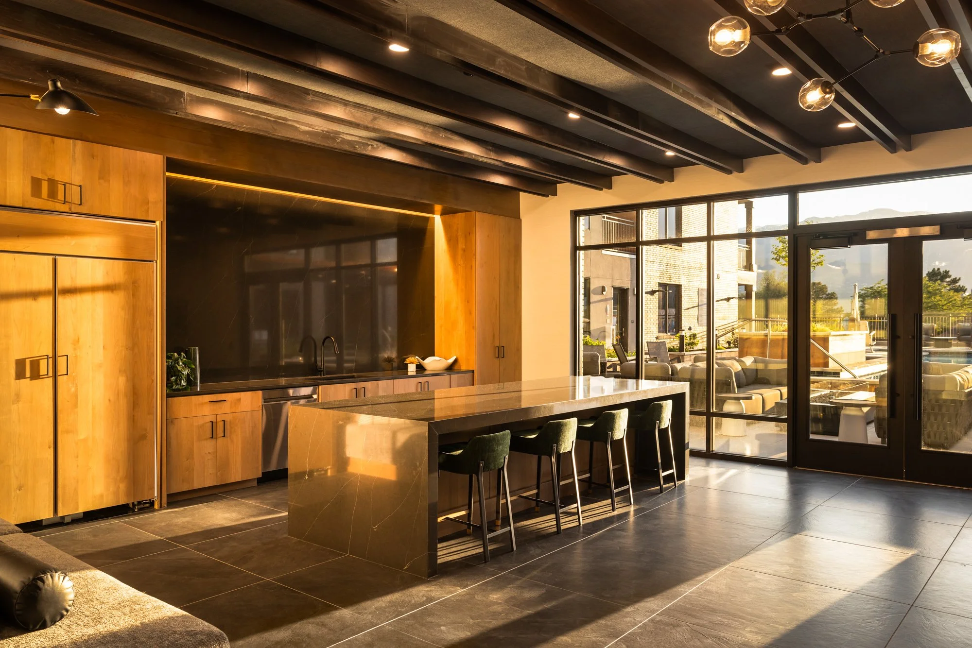 Modern kitchen with wooden cabinets, a marble island with four chairs, black ceiling beams, and large glass sliding doors opening to an outdoor seating area with a pool and mountains in the background.