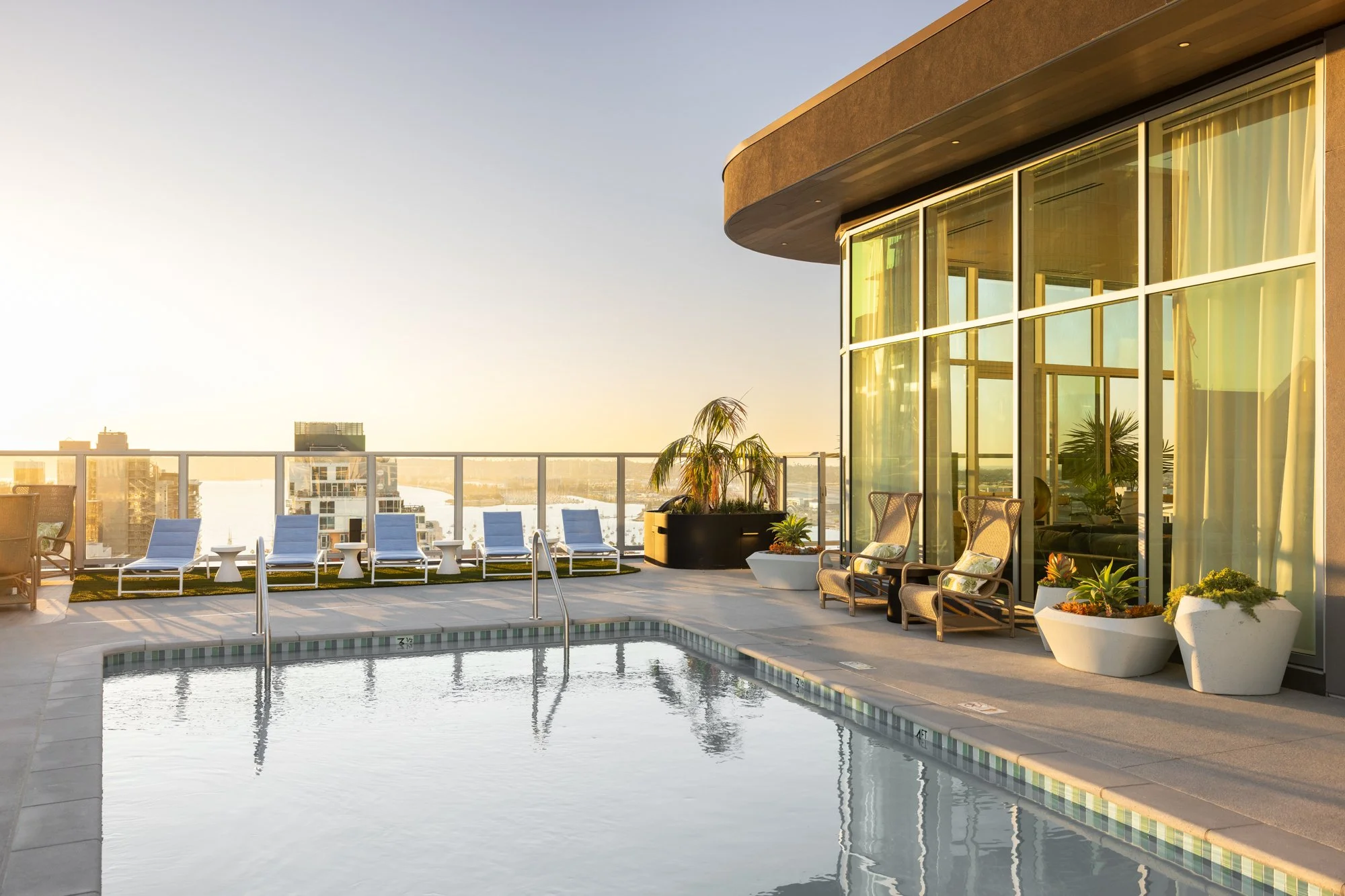 City rooftop pool area with lounge chairs, potted plants, and a glass building exterior during sunset.