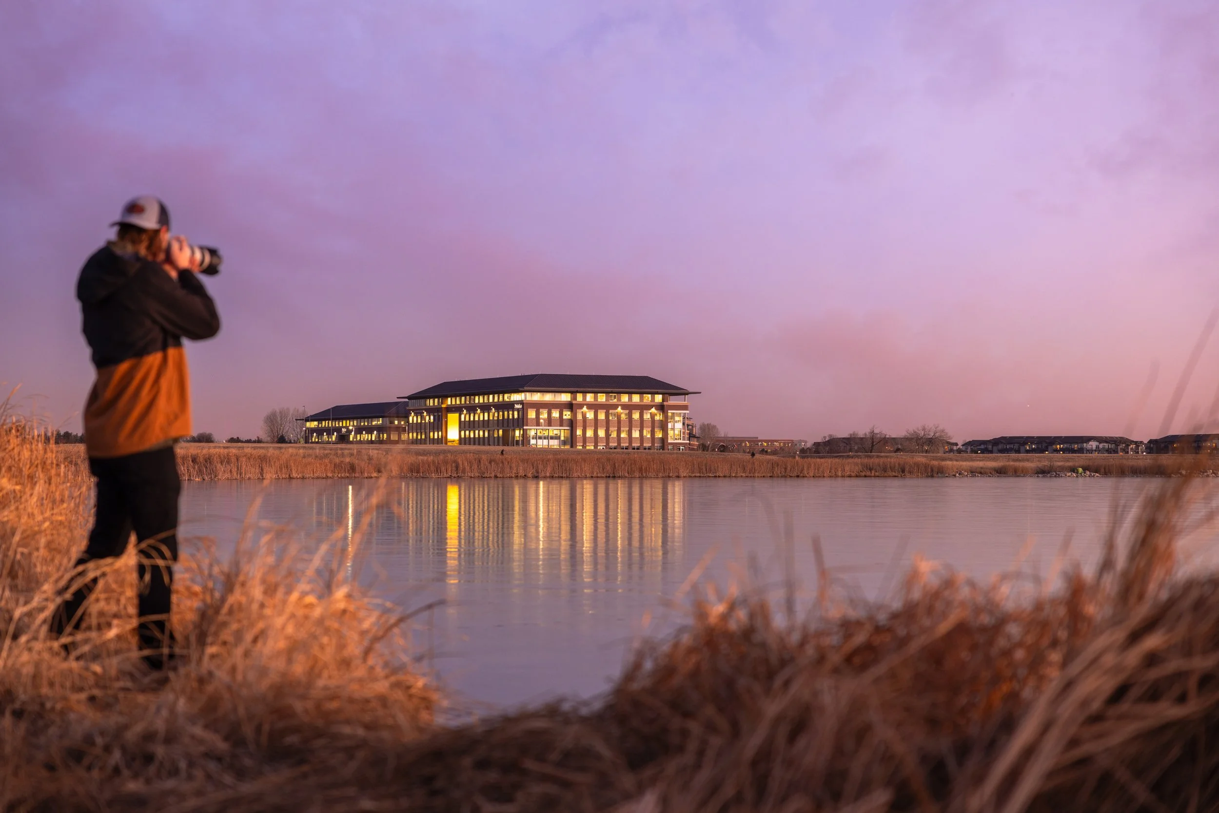 A photographer captures a large, illuminated building at sunset, reflected in a calm body of water, surrounded by dry grasses.