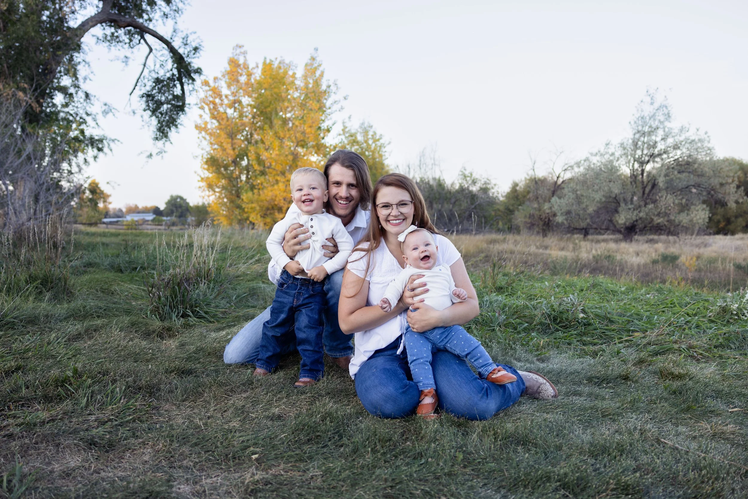Family with two children sitting on grass in outdoor setting with trees in background.