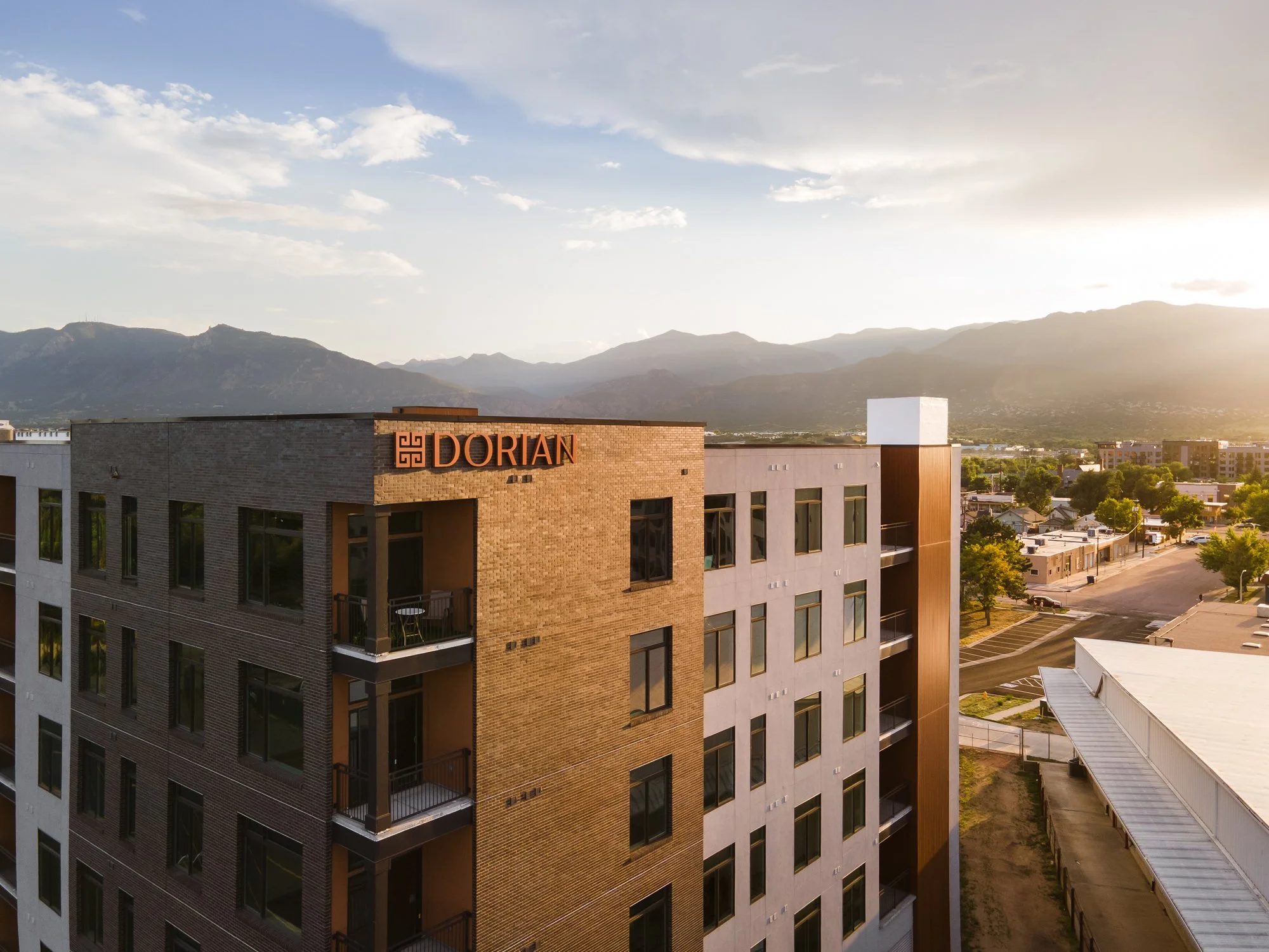 A high-rise apartment building with the sign 'Dorian' on the top, overlooking a suburban area with trees and other buildings, mountains in the background, and a sunset or sunrise sky.