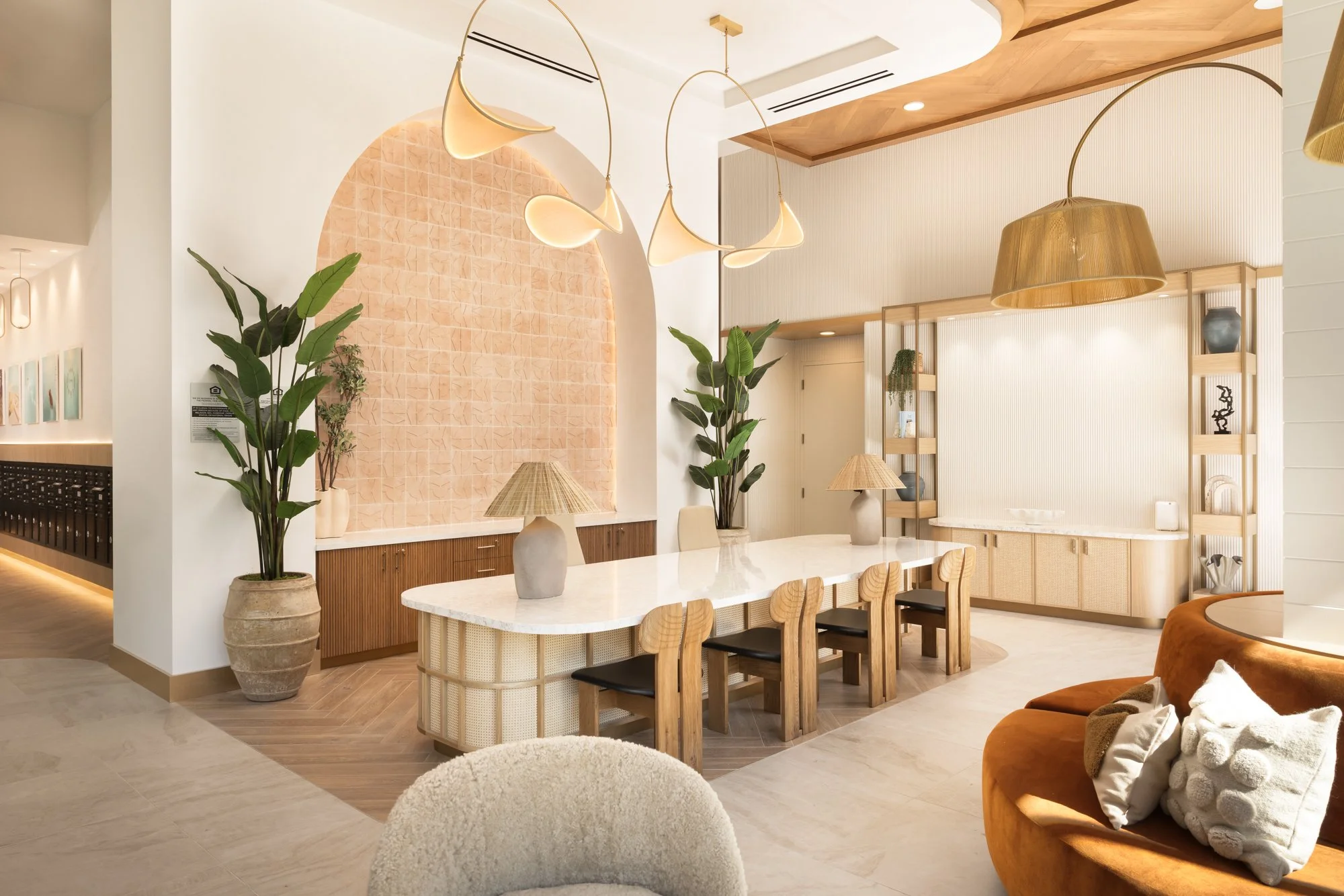 Modern interior of a dining area with a long white marble table, six wooden chairs with black cushions, large indoor plants, beige decorative lamps, and warm lighting.