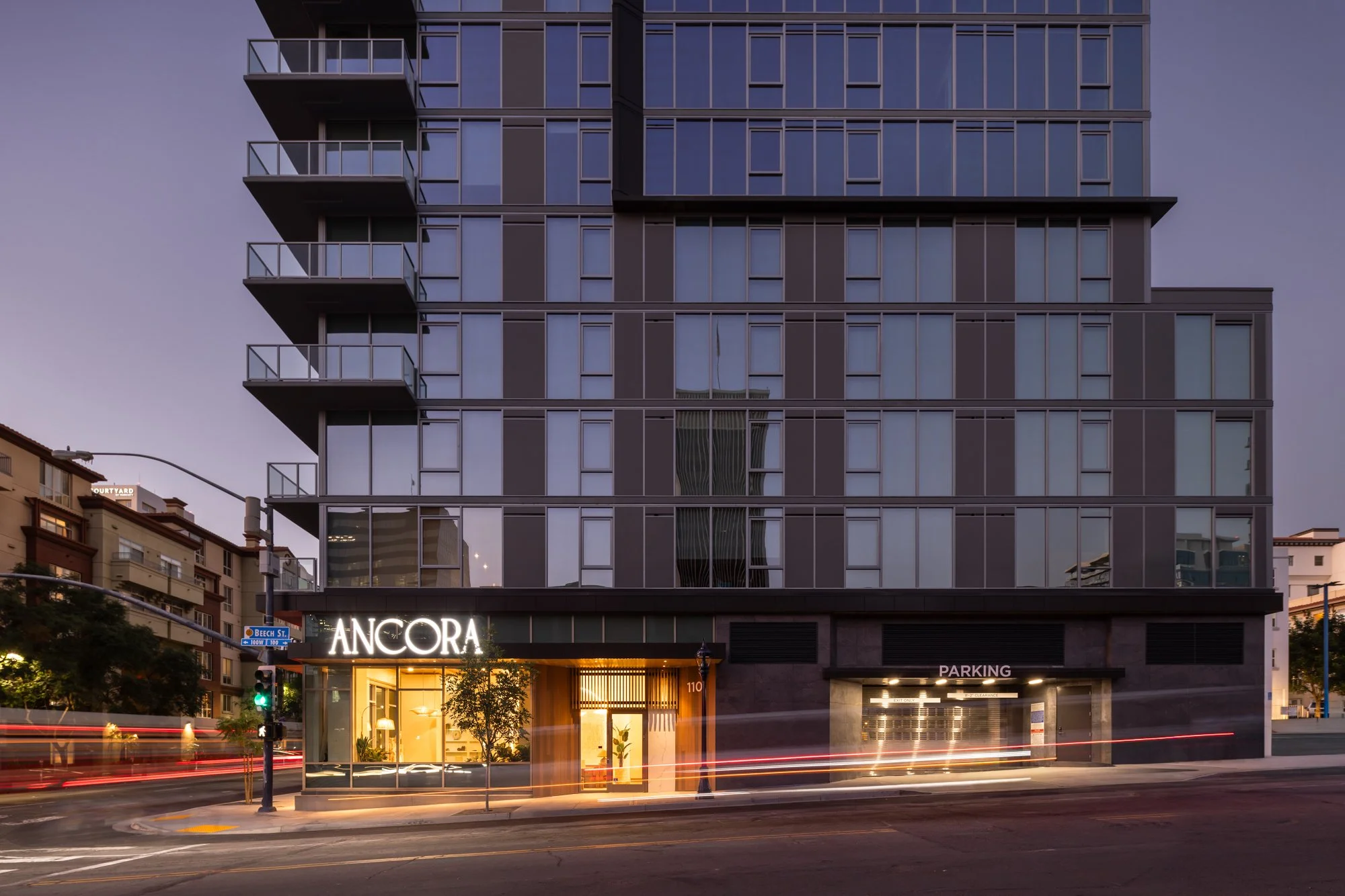 Modern multi-story building with glass facade, illuminated signage reading "ANCORA," and parking garage entrance at street level, during dusk.