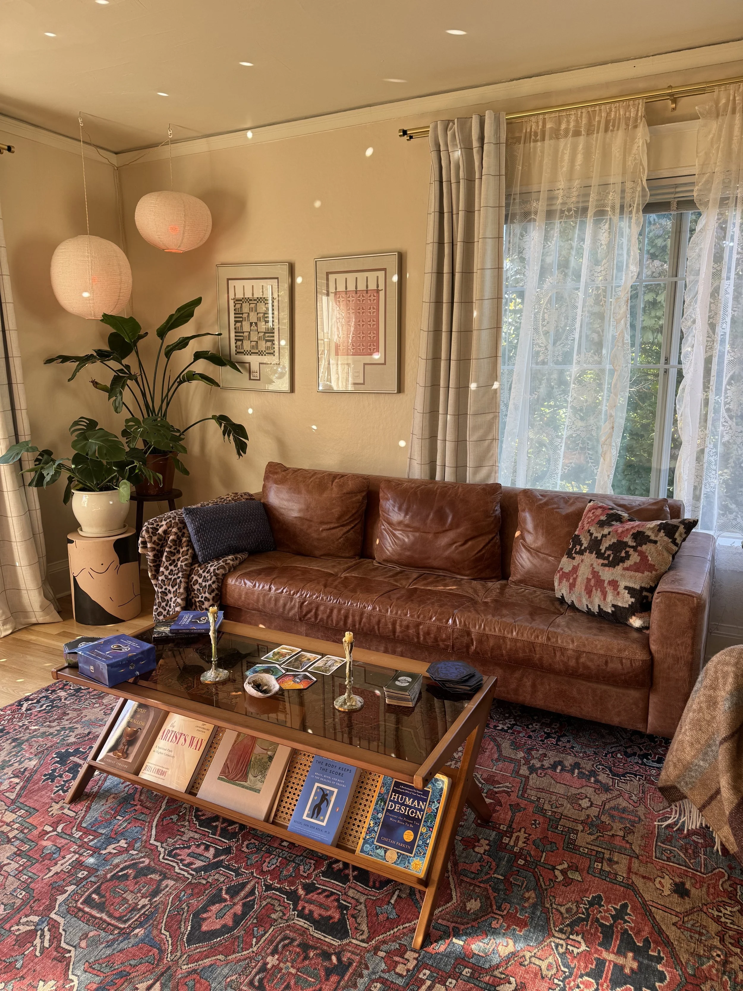 A cozy living room with a brown leather sofa, a wooden coffee table with books and tarot cards, a large leafy green plant in a decorative pot, framed artwork on the wall, lace curtains and beige drapes on the window, and hanging paper lanterns.