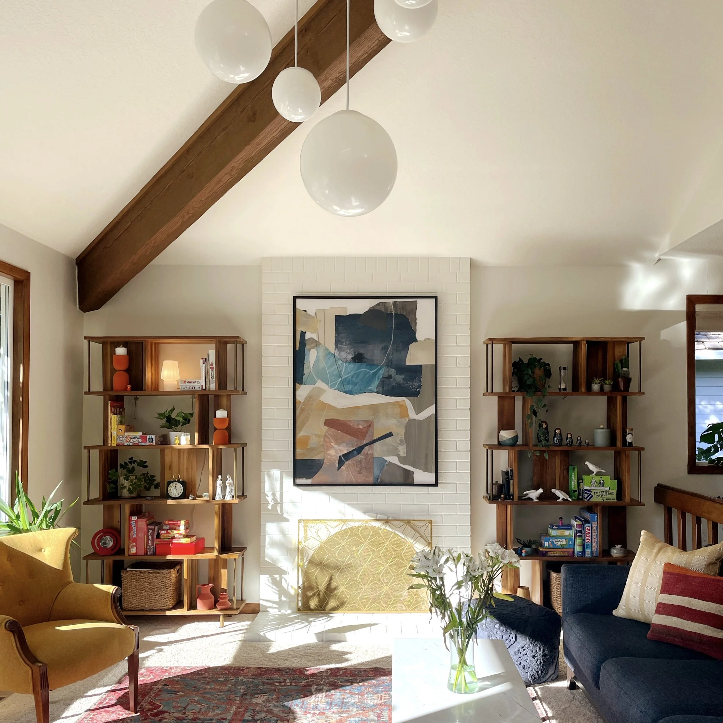 Living room with white brick fireplace, abstract art above it, wooden ceiling beams, and two wooden shelves with decor and books. Furniture includes a yellow armchair, a dark blue sofa with striped pillow, and a glass coffee table with a vase of flow