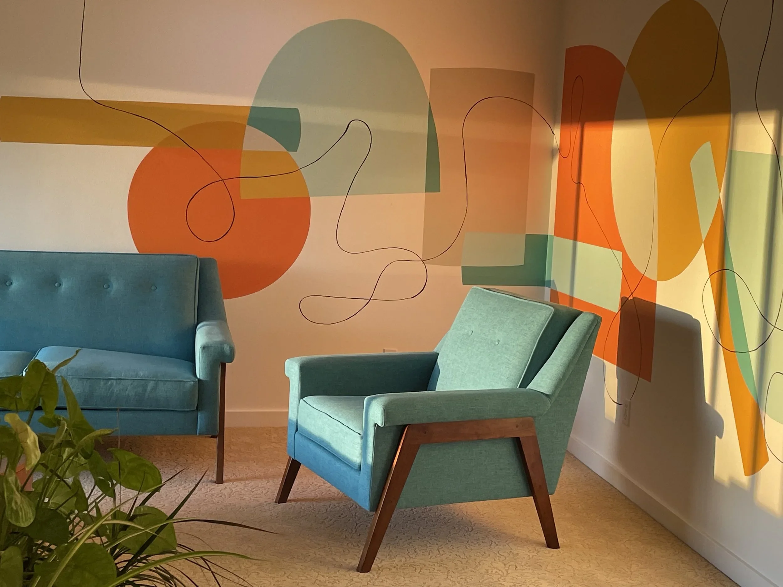 A room with abstract geometric wall art in orange, green, and blue. There are two teal-colored mid-century modern chairs and a teal sofa with a small potted plant in the foreground. The setting appears to be a waiting area or lounge.