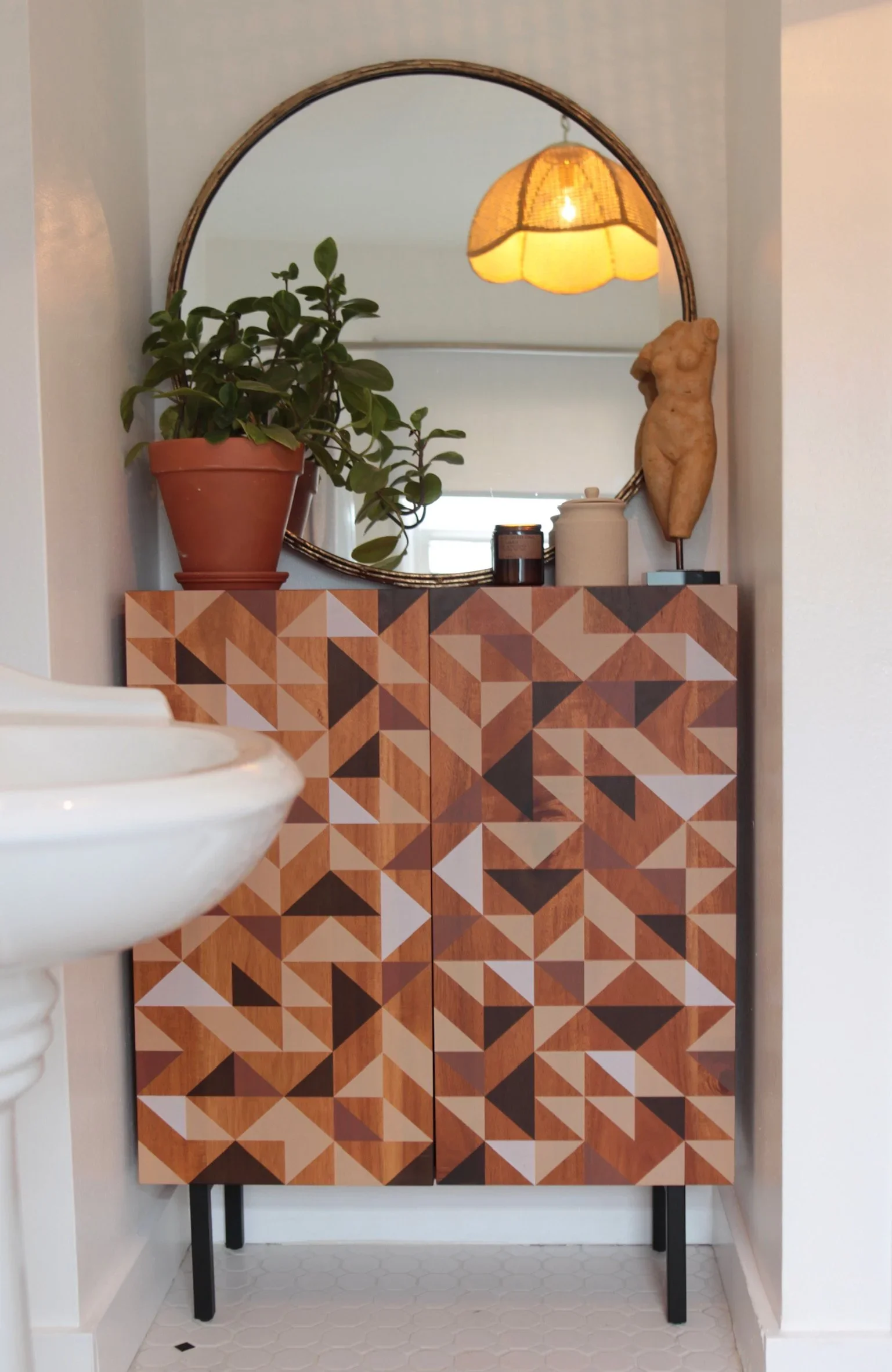 A geometric-patterned wooden cabinet with black metal legs, topped with a potted plant, candles, and a decorative torso sculpture, arranged in front of a round mirror with a wall-mounted light fixture reflected in it.