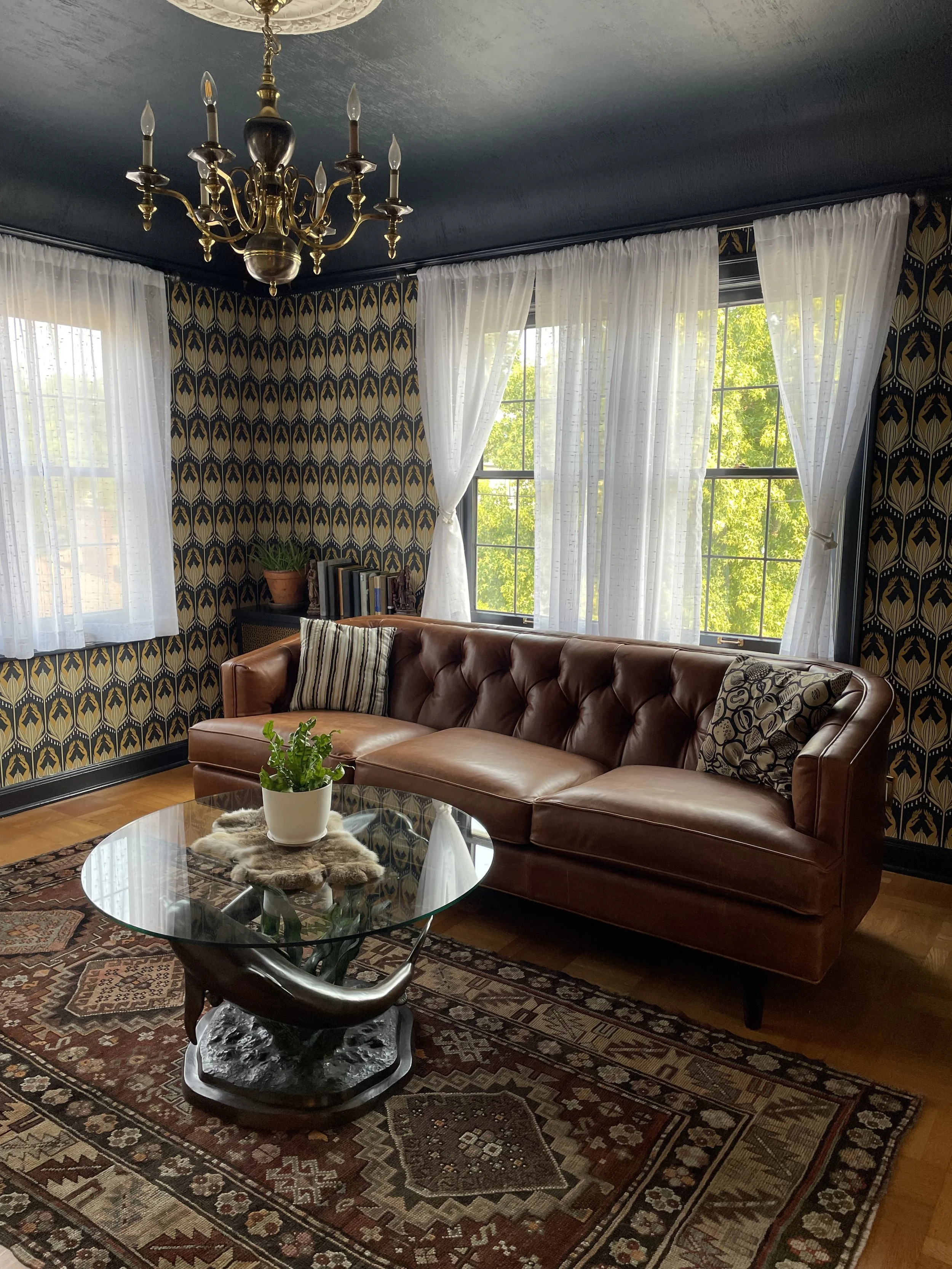 A vintage living room with a leather sofa, patterned wallpaper, large windows with white curtains, a glass coffee table with a potted plant, and a chandelier.