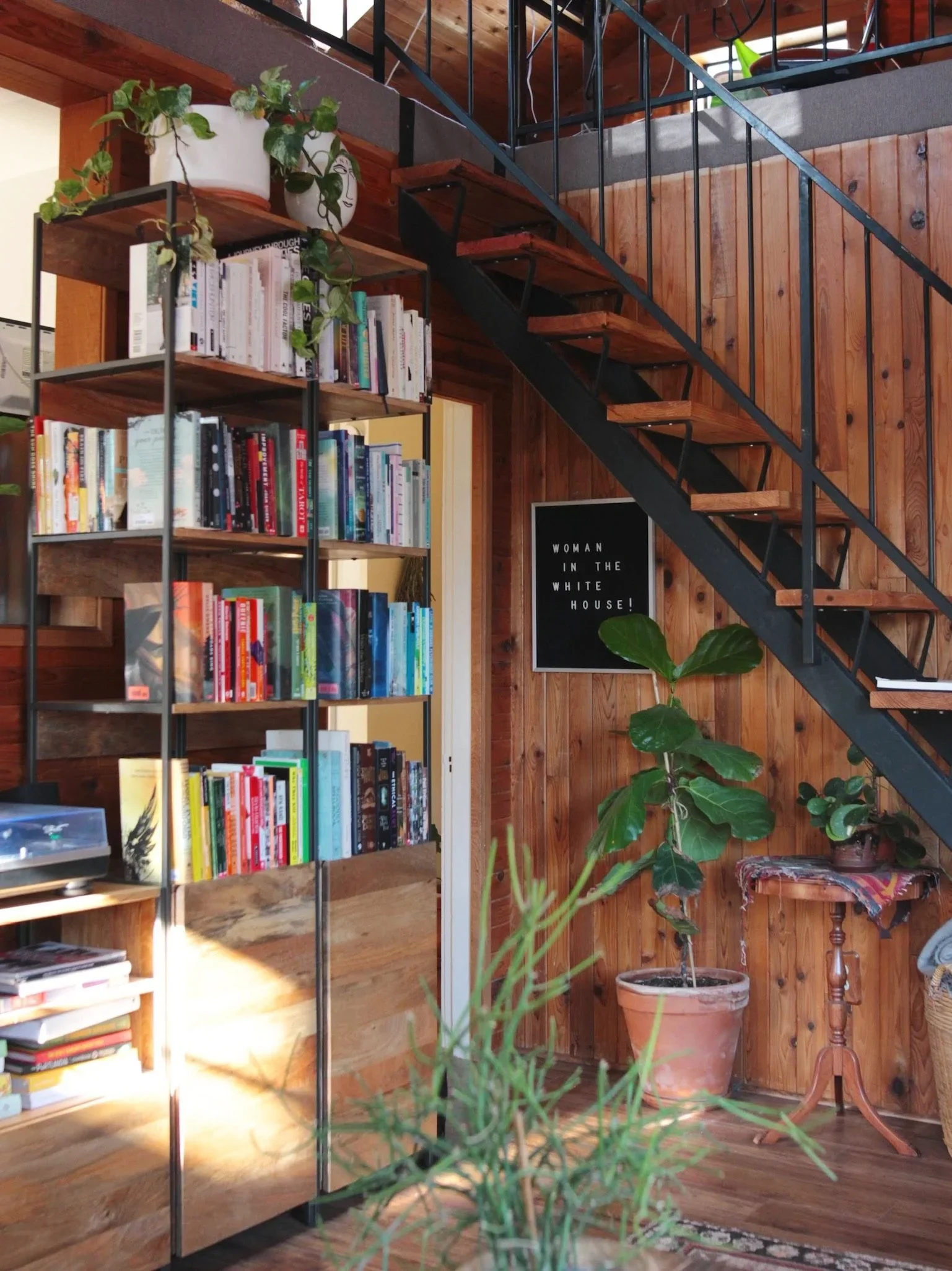 Interior of a cozy space with a wooden bookshelf filled with books, potted plants on top, and a staircase with black metal railing leading upstairs. There is a wall-mounted black poster with white text that reads 'WOMAN IN THE WHITE HOUSE!' and a sma