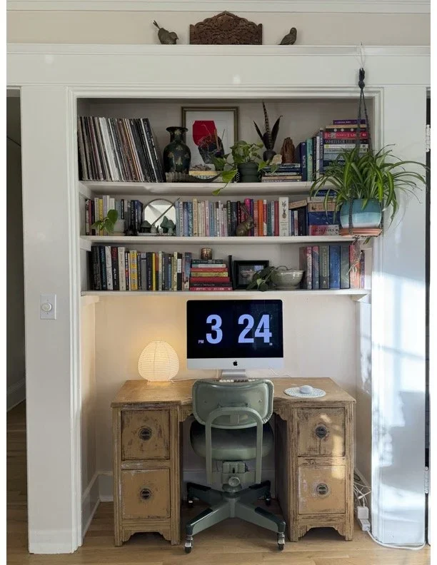 Home office setup with a vintage wooden desk, an iMac computer displaying the time 3:24, a desk lamp, and a green office chair. Behind the desk is a built-in bookshelf filled with books, plants, and decorative items. There is a window on the right le