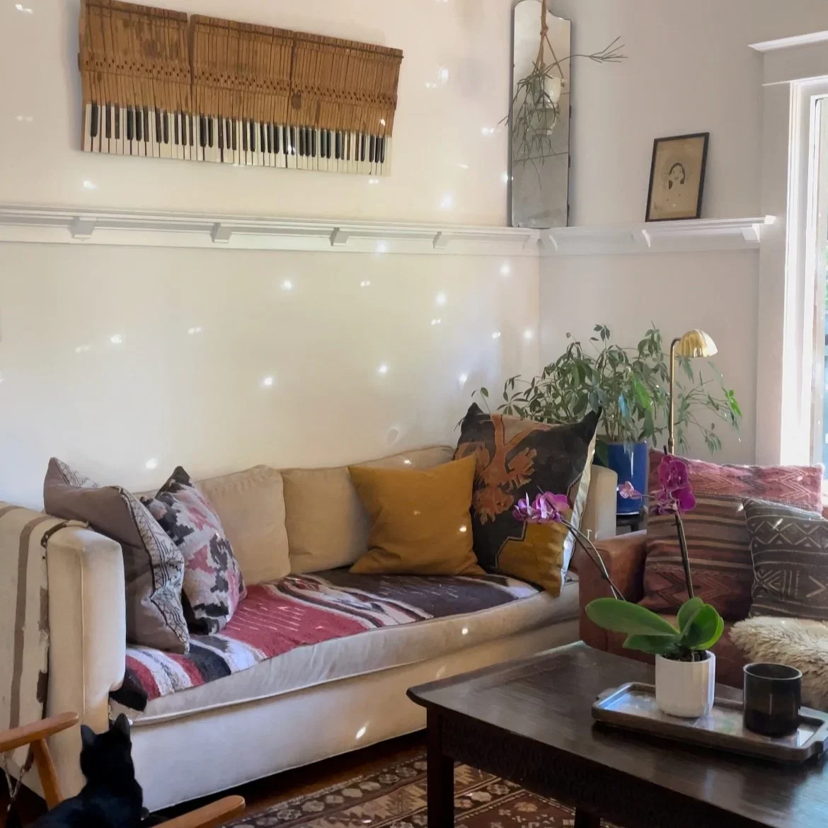 Living room with a beige sofa, colorful pillows, a wooden coffee table with a potted orchid, a lamp, potted plants, and a wall art piece resembling piano keys.