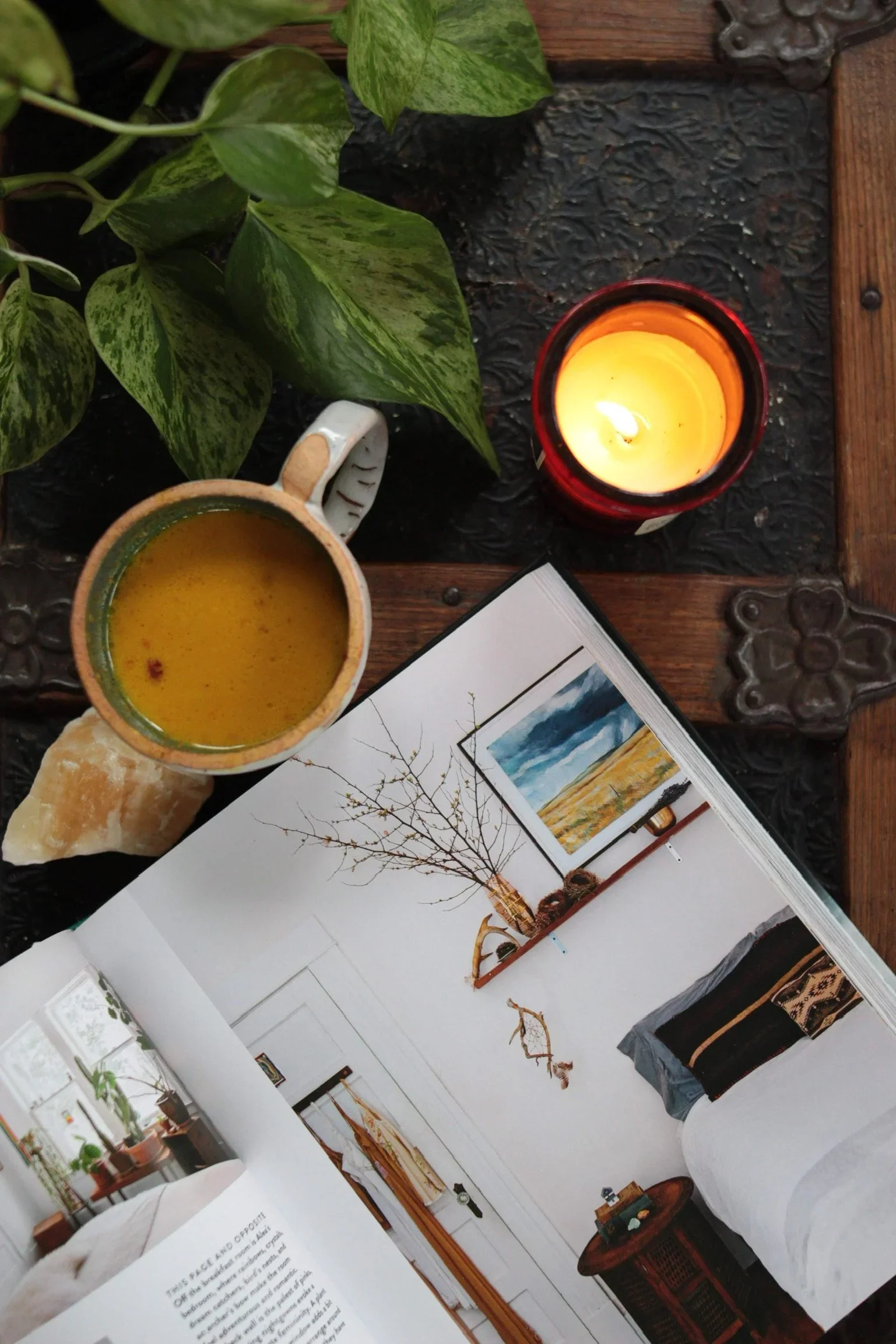 A cozy table setting with a cup of tea or coffee, a lit candle in a red glass holder, an open magazine featuring home decor, and some green leaves in the background.