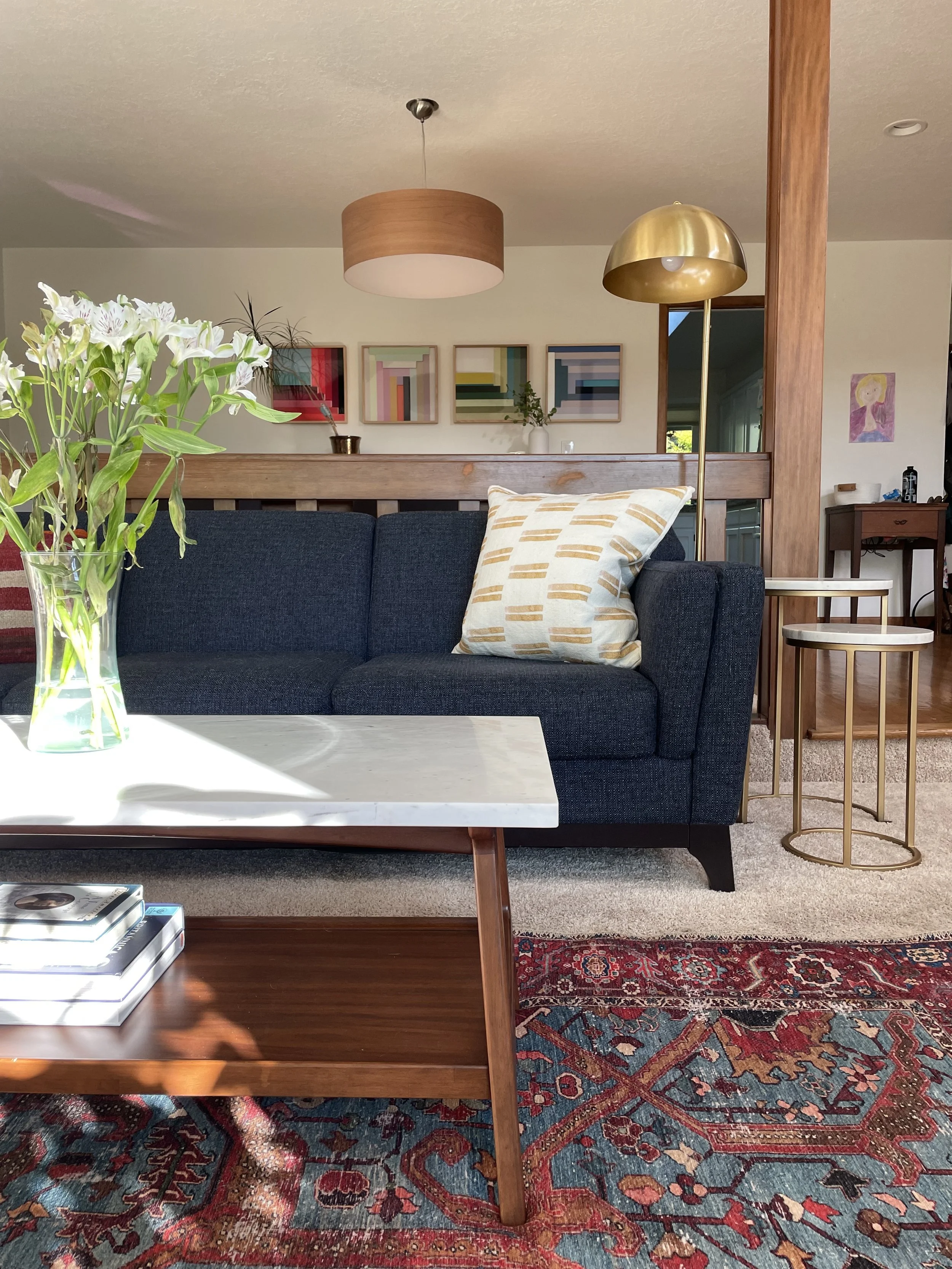 Living room with a dark blue sofa, white marble coffee table, floral pillow, lamp, artwork, and wooden accents.