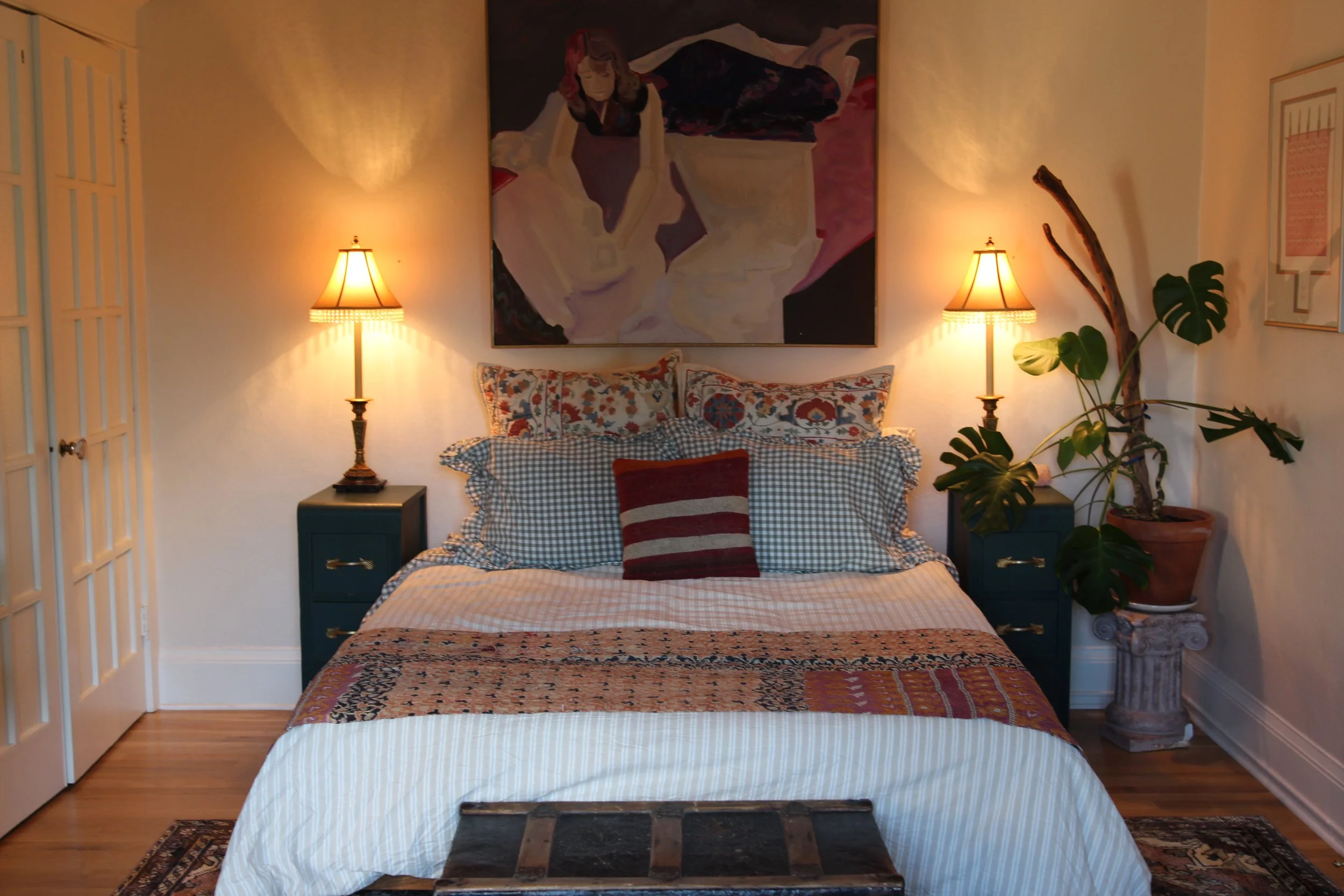A cozy bedroom with a bed decorated with patterned pillows and a striped throw blanket. Two matching lamps on dark blue nightstands are on either side of the bed. A large framed abstract painting hangs above the bed, and a potted monstera plant is on