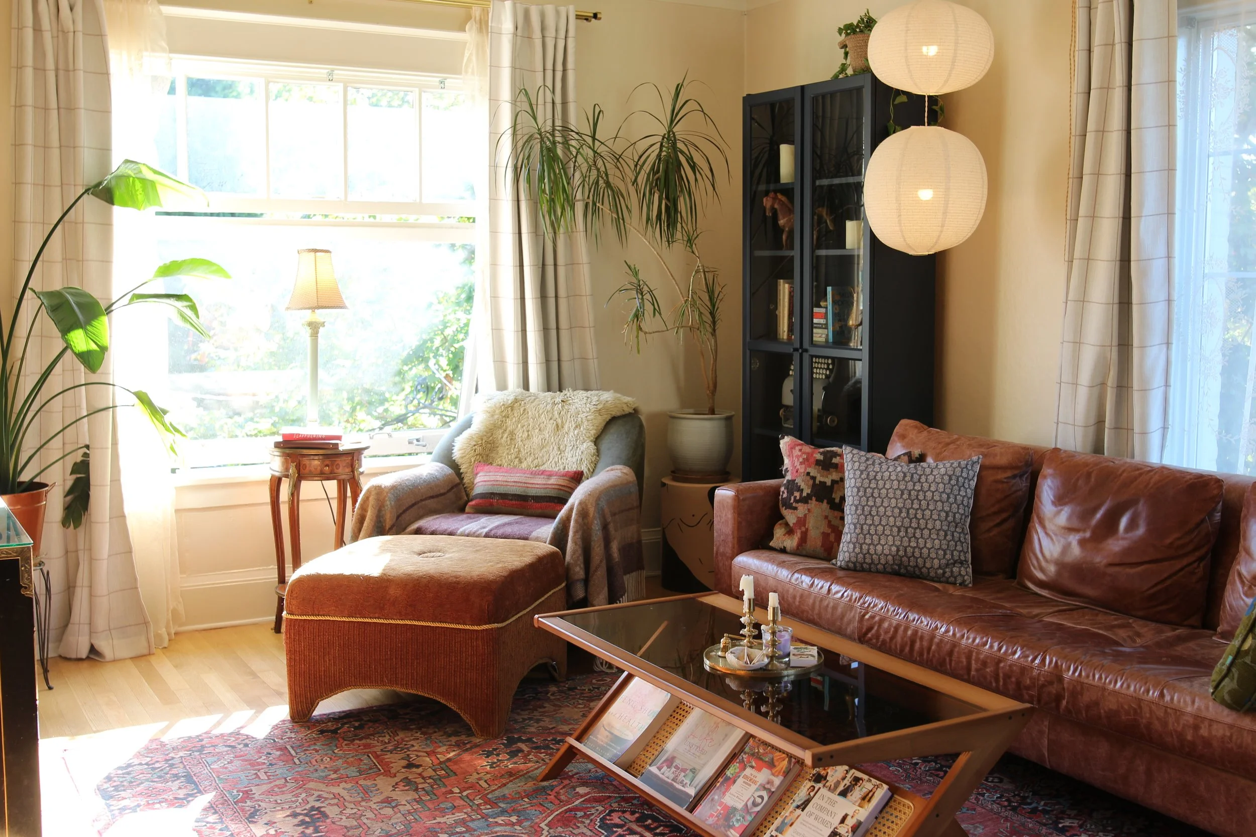 Living room with large window, armchair with throw blanket, leather sofa with patterned pillows, wooden coffee table with magazines, bookshelf, potted plants, and hanging paper lanterns.
