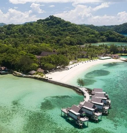 Overwater bungalows on a tropical beach with white sand and clear turquoise water, surrounded by lush green hills.