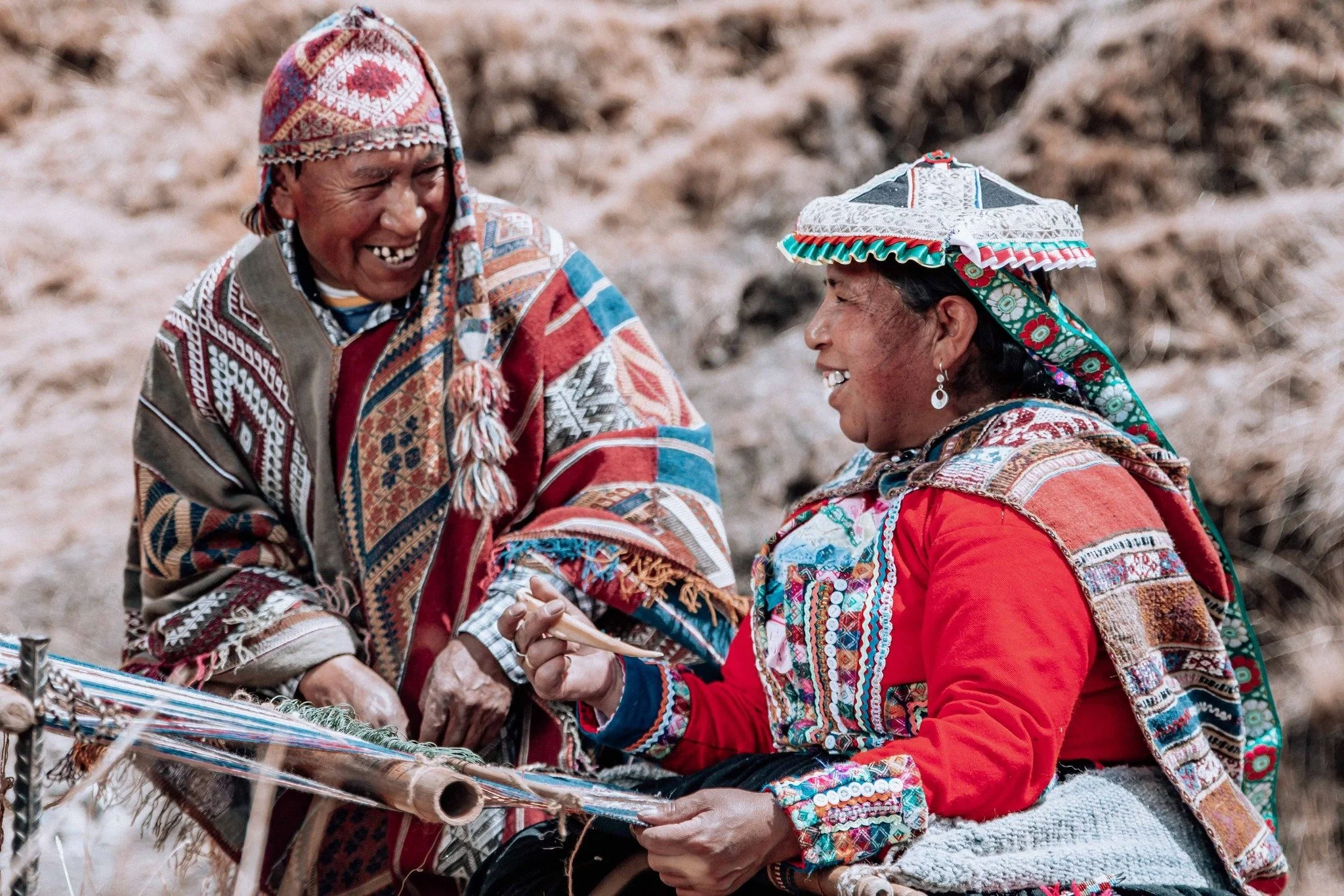 Two older women in traditional colorful clothing and headgear, smiling and holding hands outdoors against a natural background.