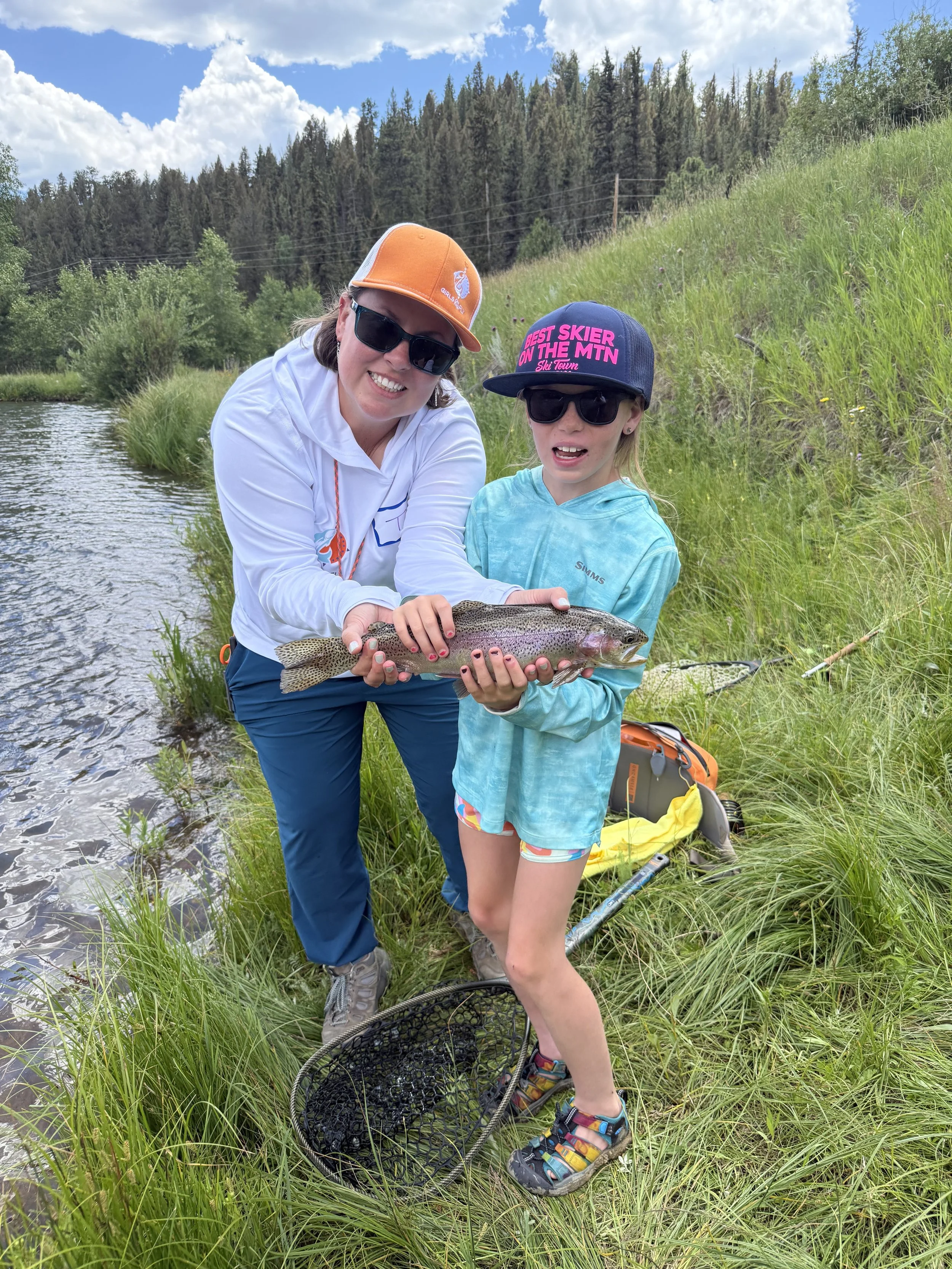 A woman and a girl holding a fish by a riverbank with grass, trees, and a sky with clouds in the background.
