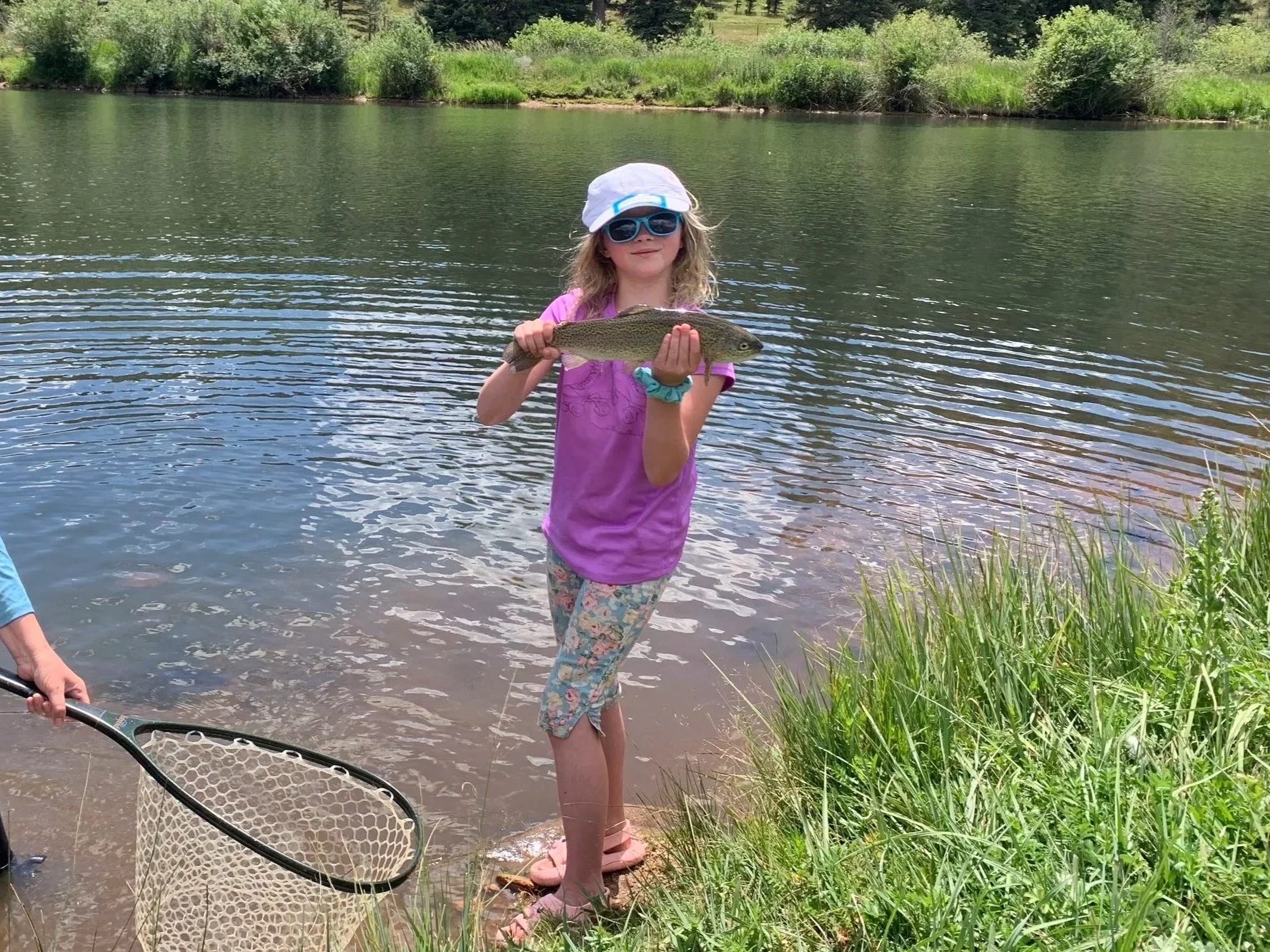 A young girl stands at the edge of a lake holding a fish. She is wearing sunglasses, a light-colored hat, a bright pink shirt, and floral-patterned leggings. A fishing net is visible in the foreground, and lush green bushes and trees are in the background.