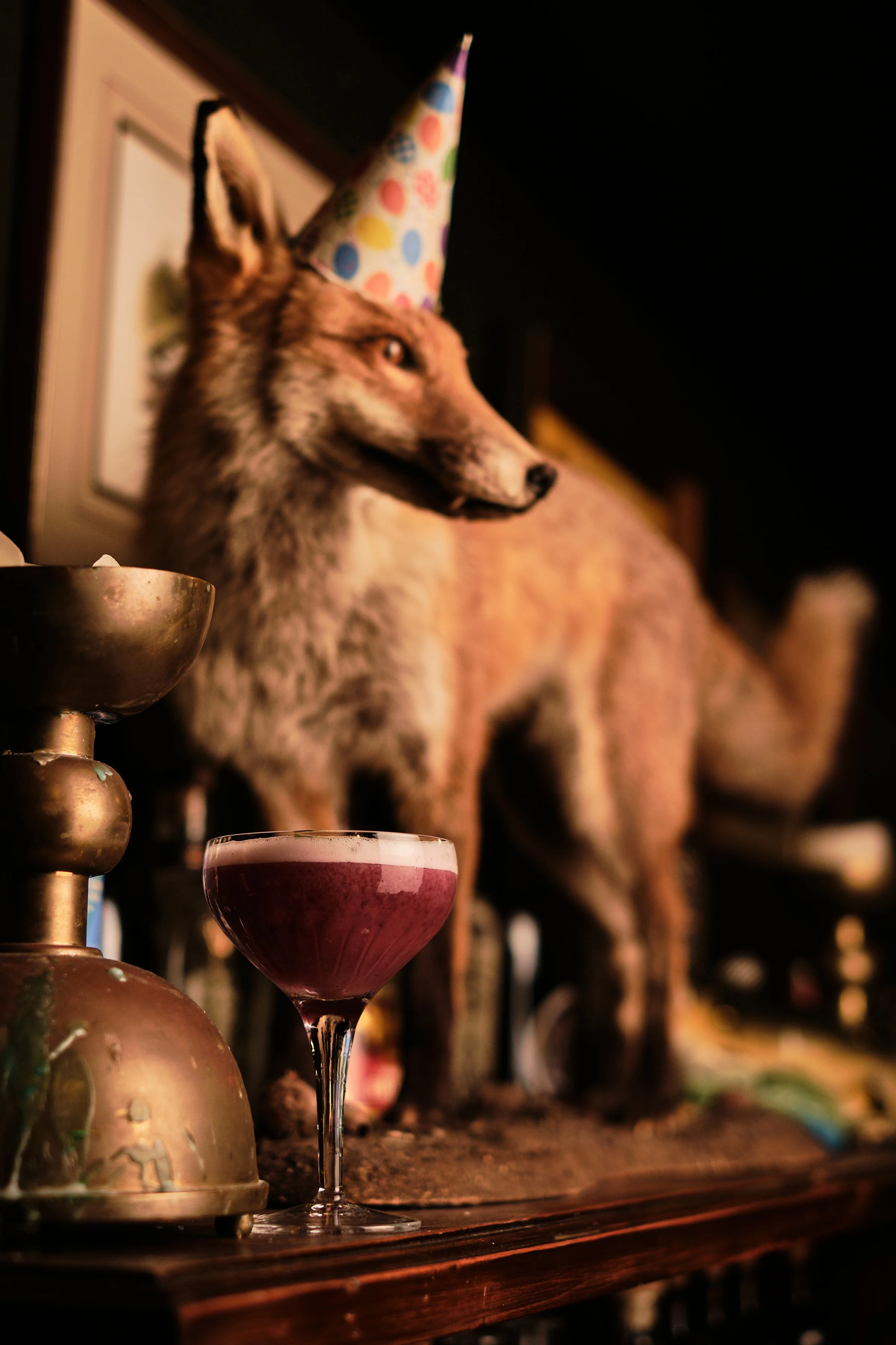 A dark purple cocktail in an elegant coupe sits on a mantle next to spirit bottles, candle holders and a taxidermy fox with a party hat on.