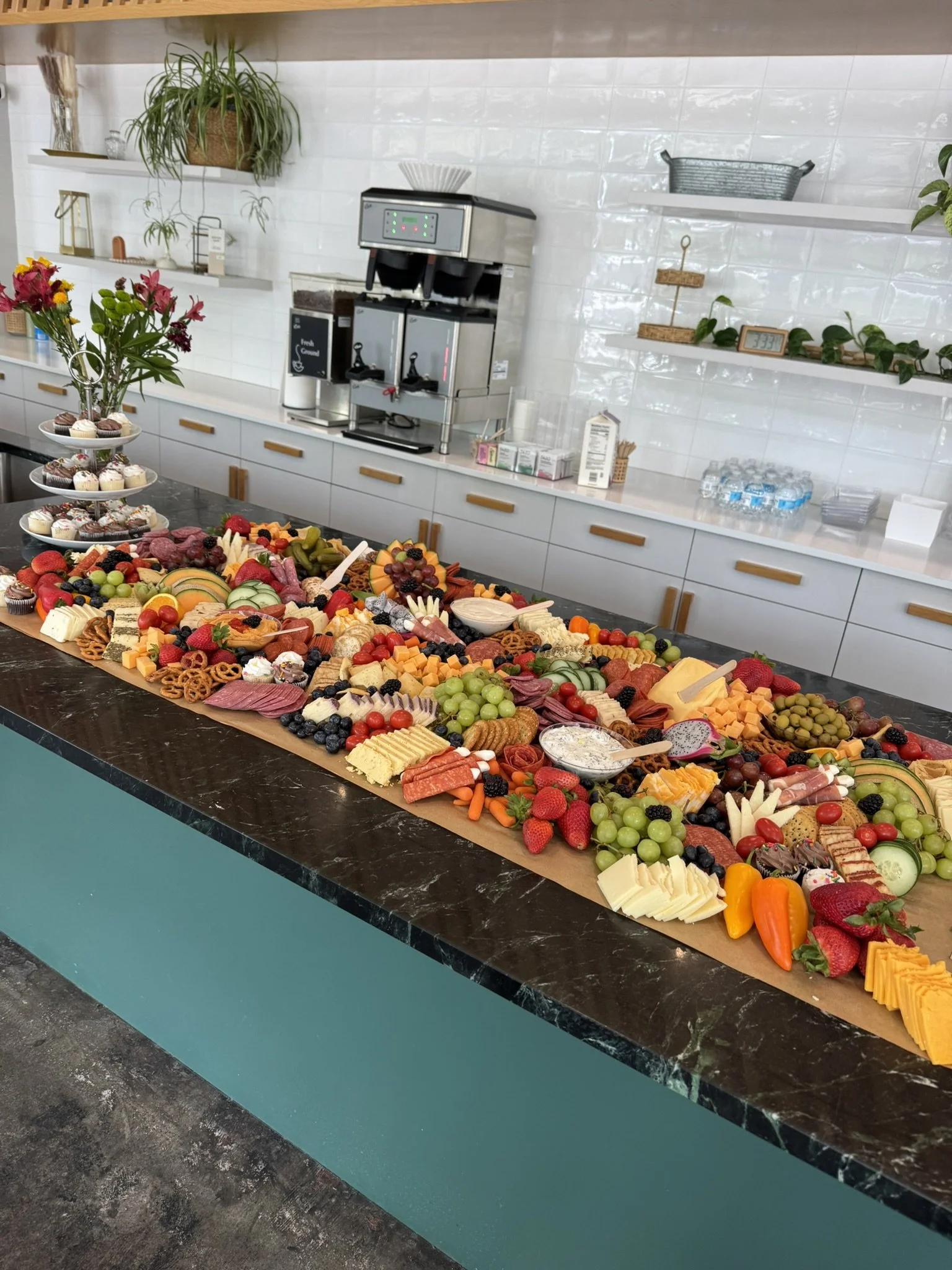 A long table with a variety of colorful fruits, cheeses, and charcuterie arranged on a brown paper covering in a modern kitchen setting.