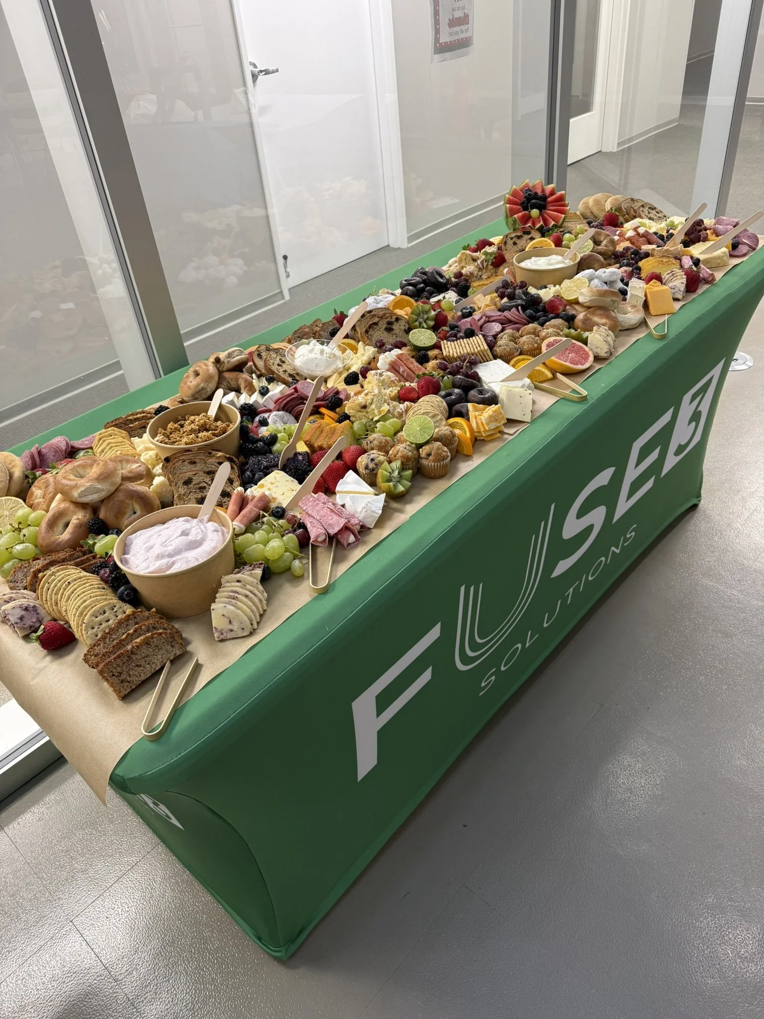 A buffet table with assorted fruits, baked goods, cheeses, and spreads, set up with serving utensils. The table has a green cover with the logo 'FOLUEED SOLUTIONS' on the side.