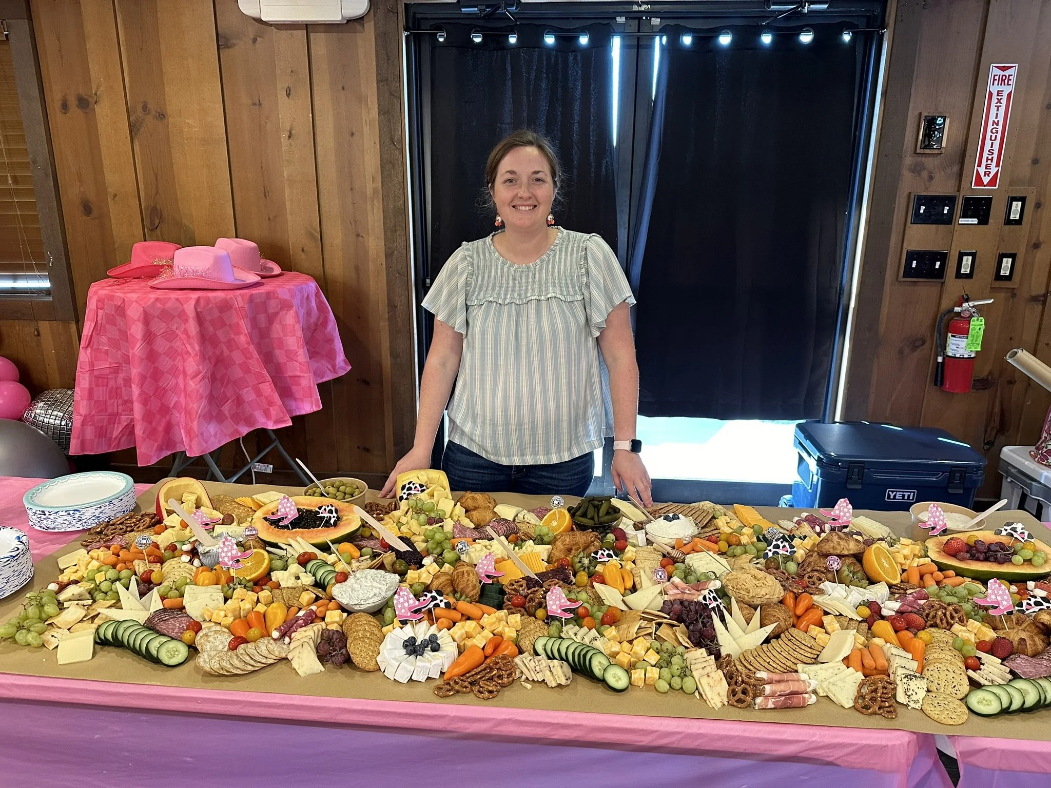 Woman standing behind a large table of cheese and fruit platter at a party, with pink decorations and cowboy hats.