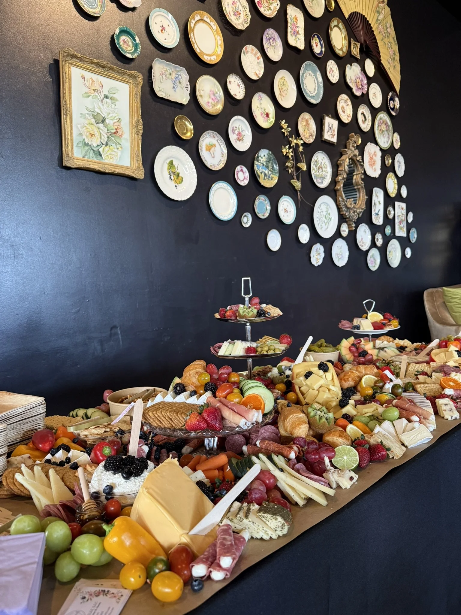 A table of assorted cheeses, fresh fruits, crackers, and various snacks with a decorative wall of plates and framed art in the background.