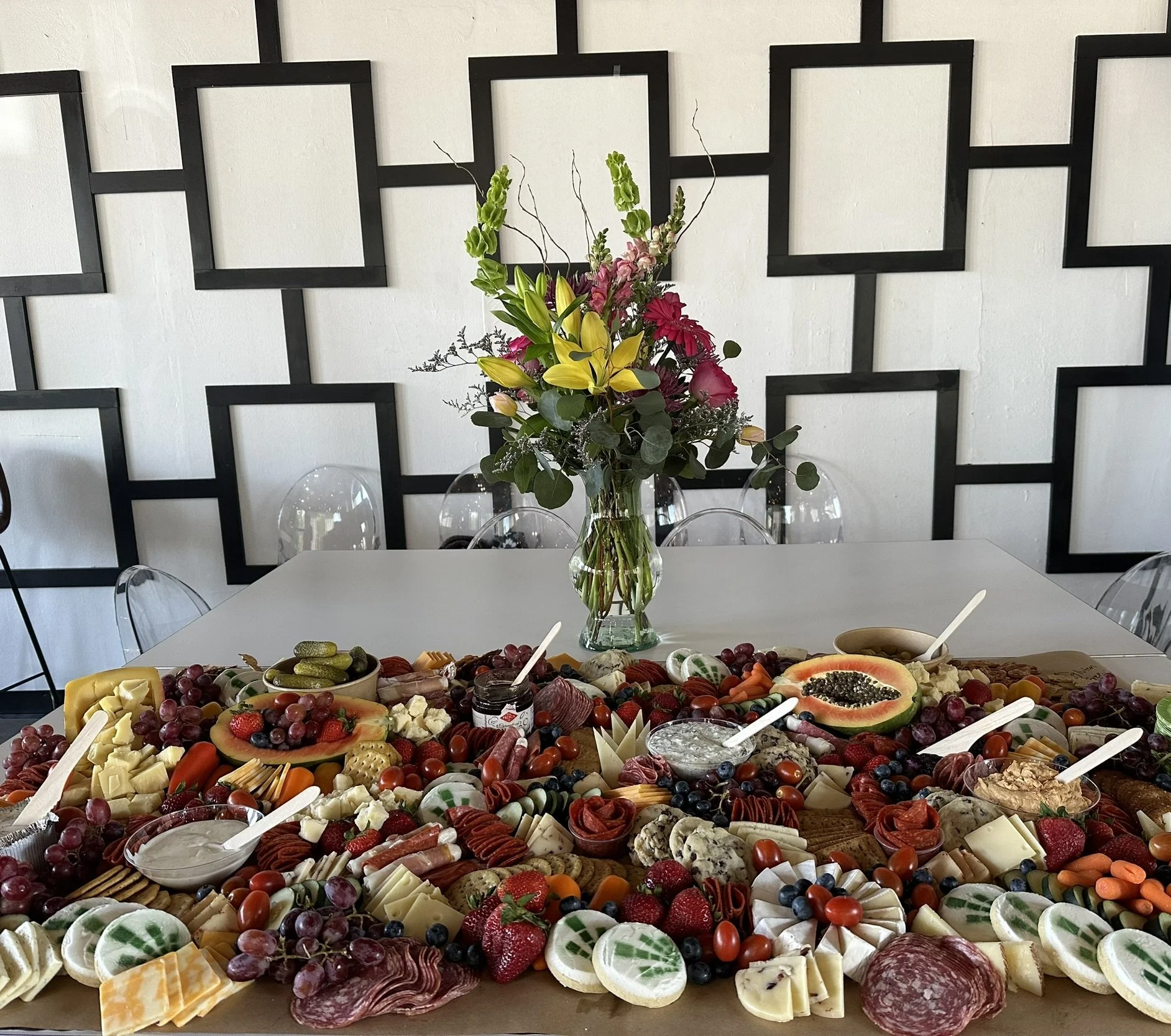 A table of assorted cheeses, fruits, and charcuterie with a flower arrangement in a glass vase on a white table against a geometric black and white wall.