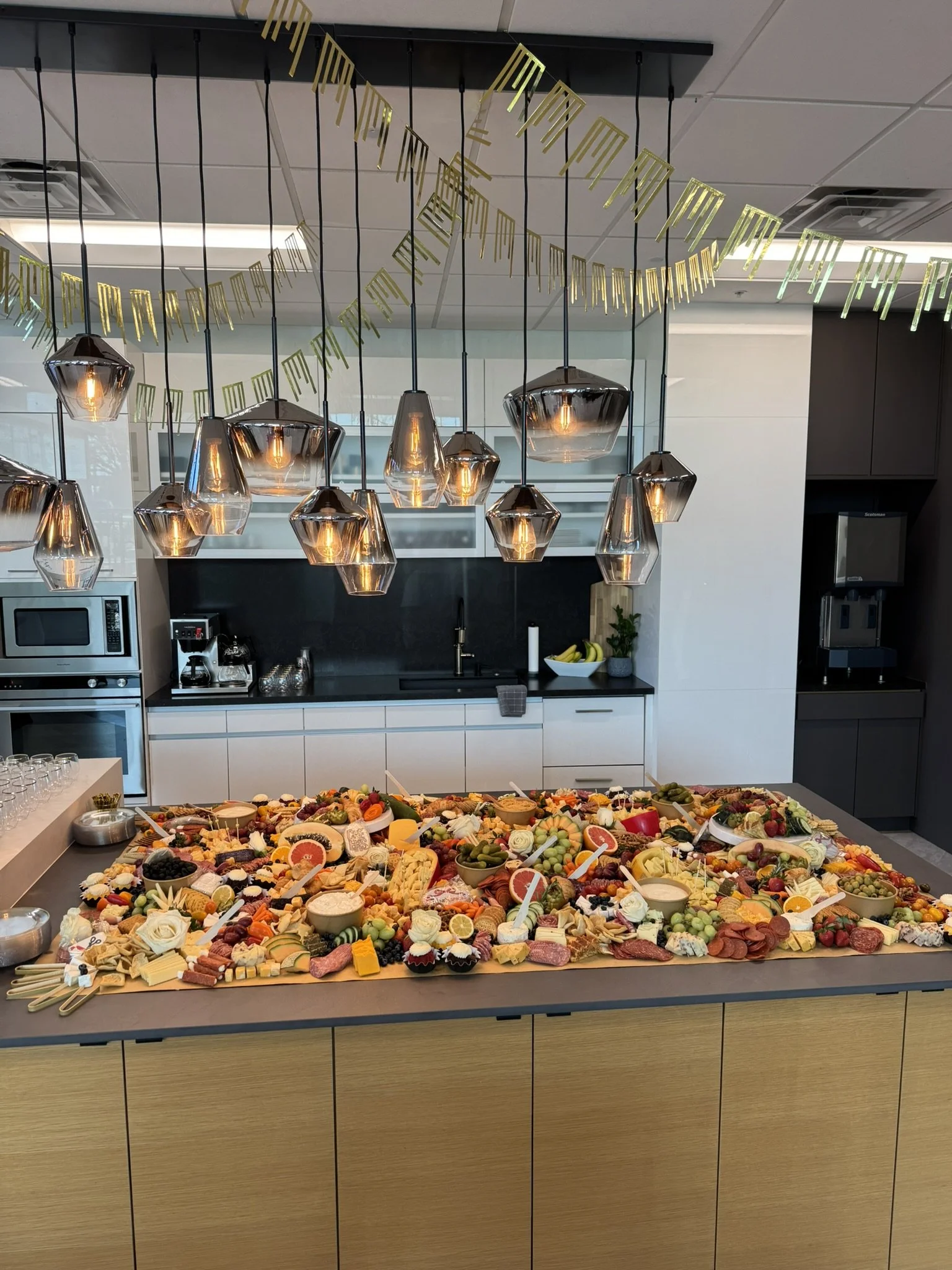 A large charcuterie board with various cheeses, meats, fruit, and appetizers on a kitchen counter decorated with gold banners and pendant lights.