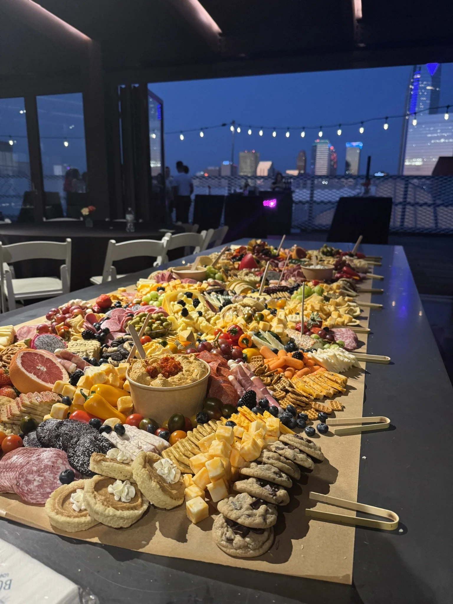 An elaborate charcuterie board with meats, cheeses, fruits, vegetables, and snacks, set on a table at an outdoor or rooftop event during dusk, with city skyline in the background.