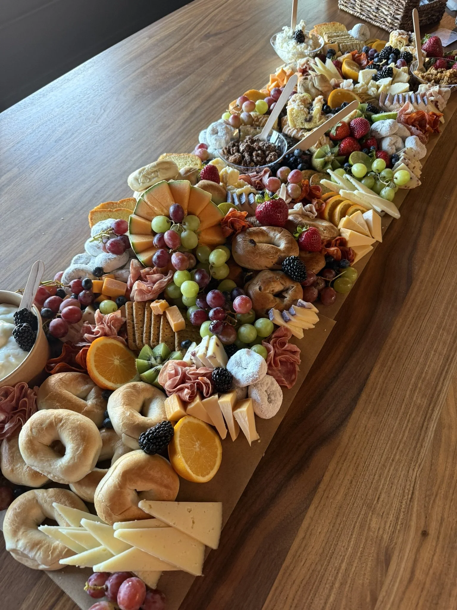 A long wooden table with a large cheese and fruit platter, including grapes, sliced oranges, cheese, crackers, bagels, and various desserts and snacks.