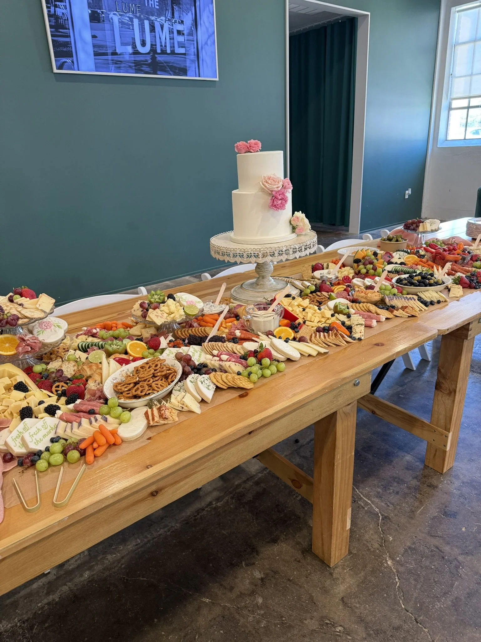 A long wooden table with a large assortment of cheeses, fruits, crackers, and vegetables, and a white tiered cake with pink and white flowers on top, set in a room with teal walls and a framed sign that says 'LUME' in the background.