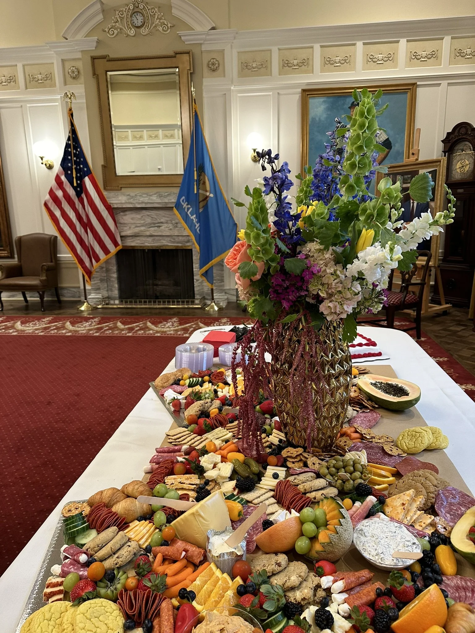 A long table with a variety of snacks, cheeses, fruits, and crackers, decorated with a large bouquet of colorful flowers in a tall, ornate gold vase. In the background, there are flags of the United States and Oklahoma, a fireplace, and framed artwor