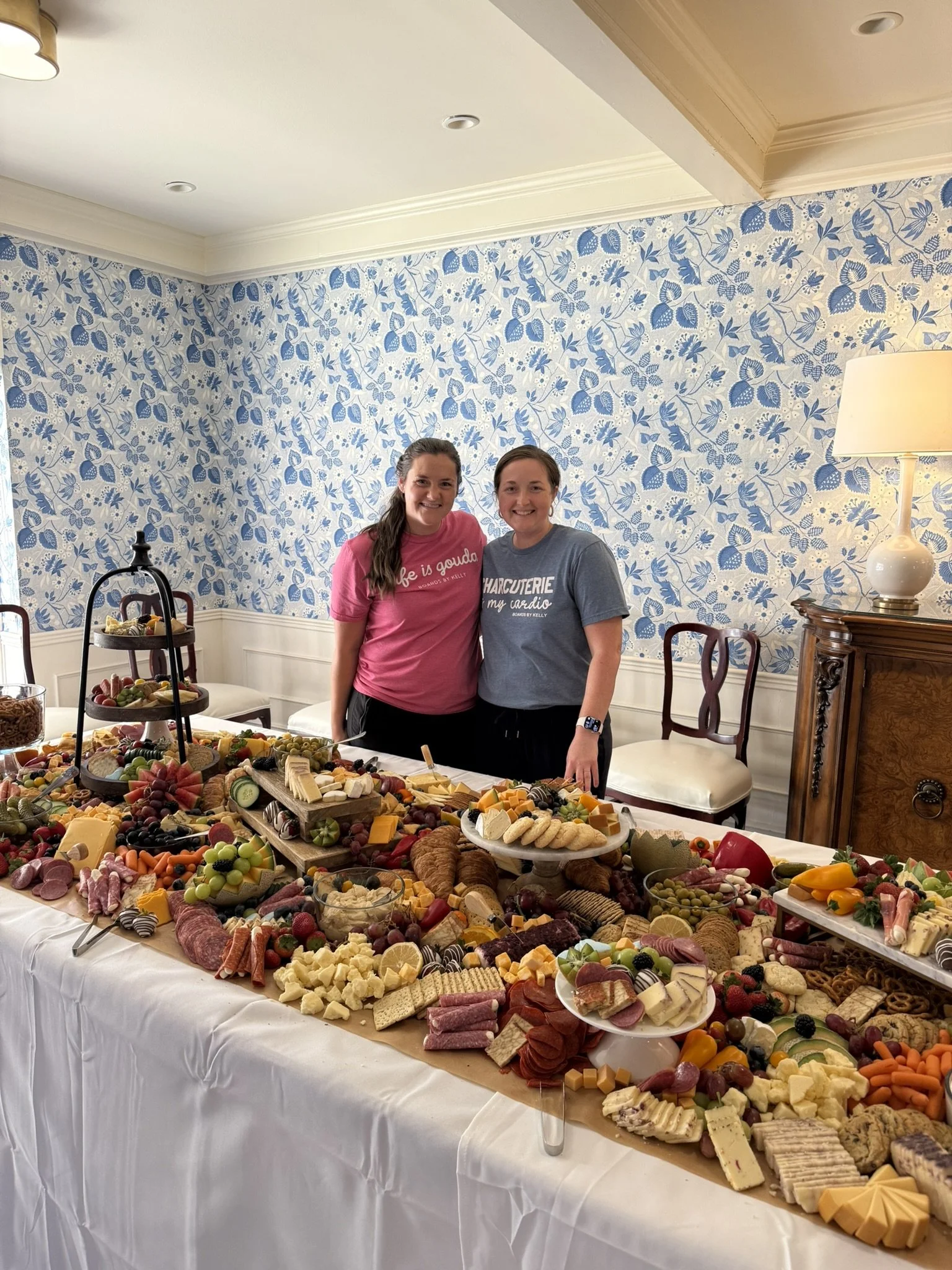 Two women standing behind a large table filled with a variety of cheeses, fruits, meats, and snacks, in a room with a floral blue and white patterned wall and a wooden dresser.