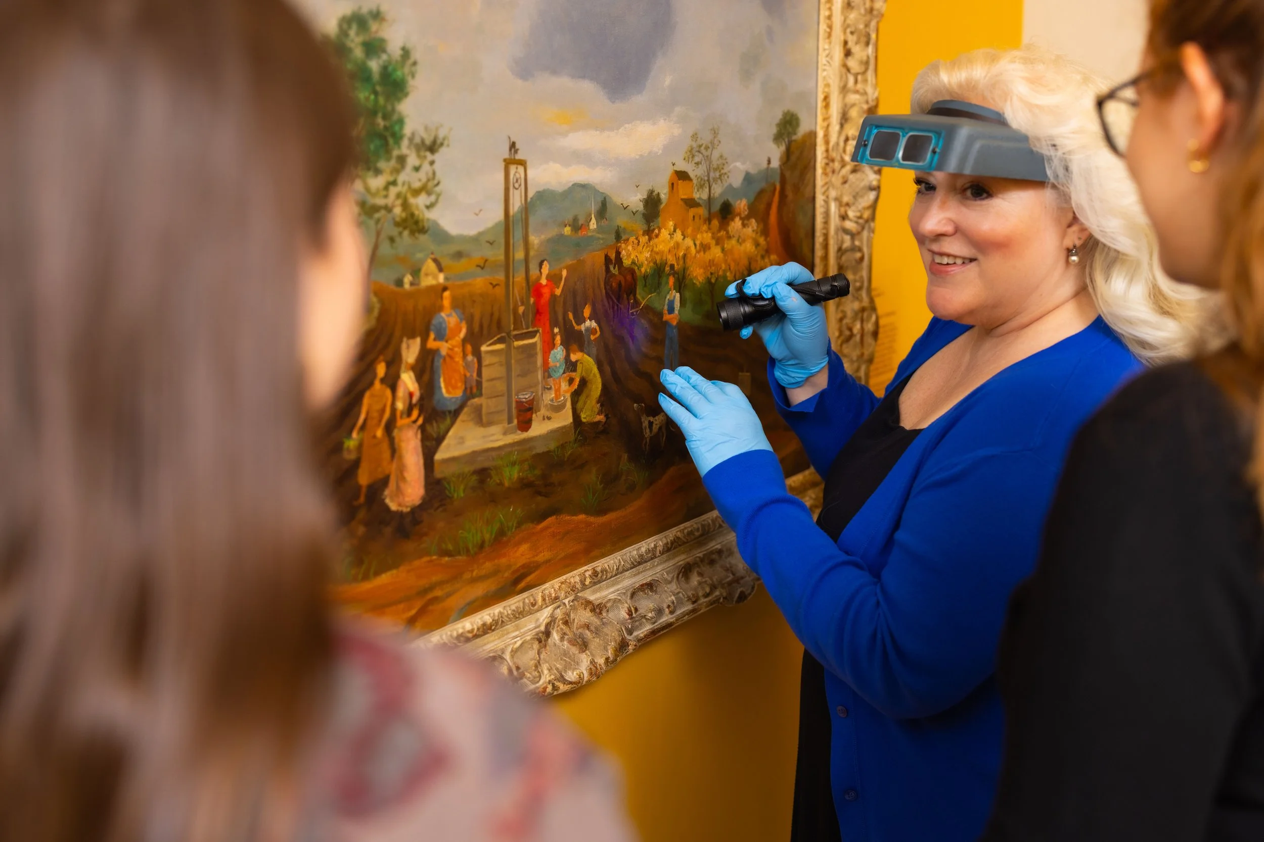 An older woman with white hair, wearing blue gloves, a magnifying loupe, and a blue cardigan, examining a landscape painting with a small group of people observing at an art gallery.