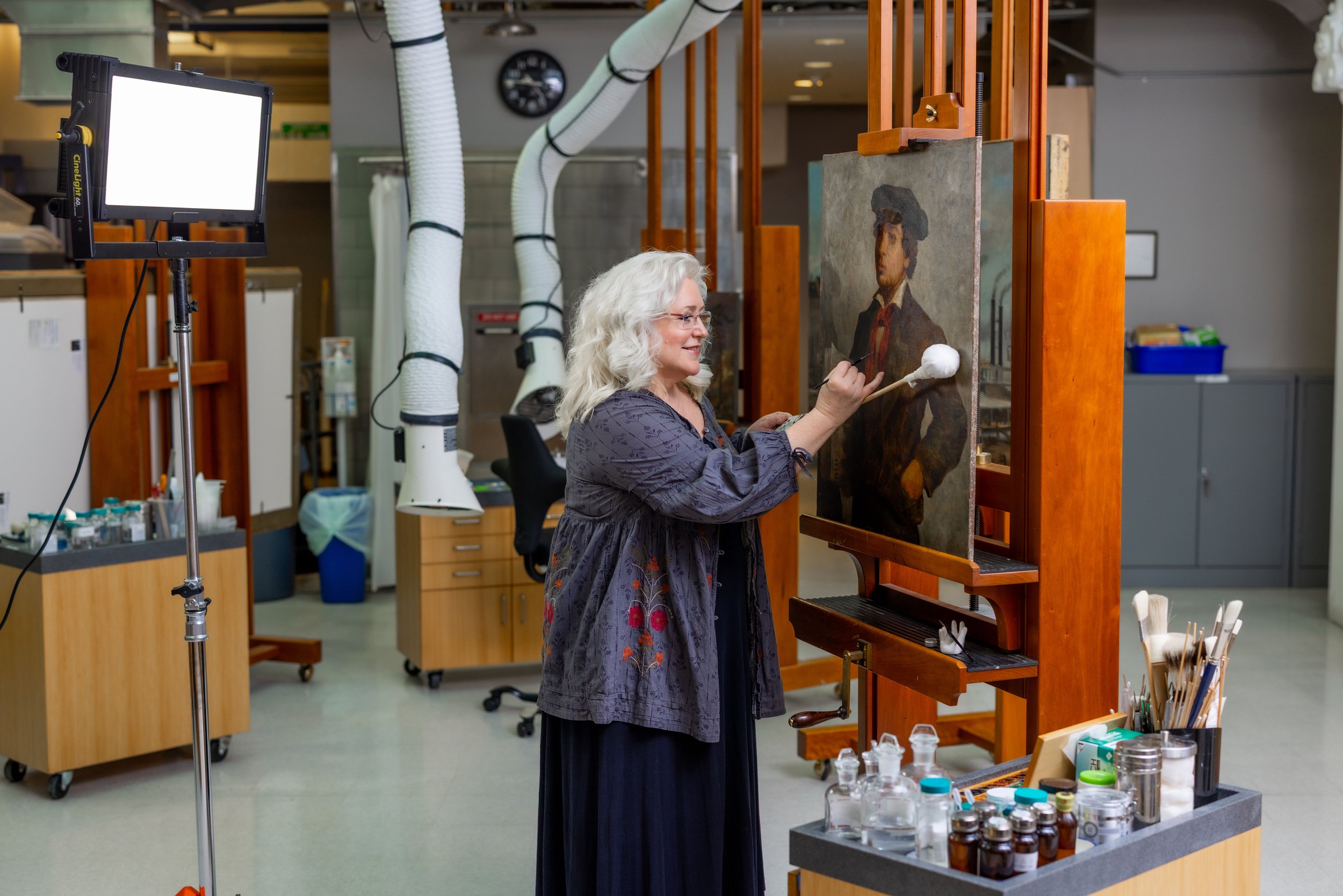 A woman with gray, curly hair paints a portrait of a woman with black hair on an easel in an art studio. The studio has various art supplies, bottles, and jars on a table, with a large soft light on a stand illuminating the scene.