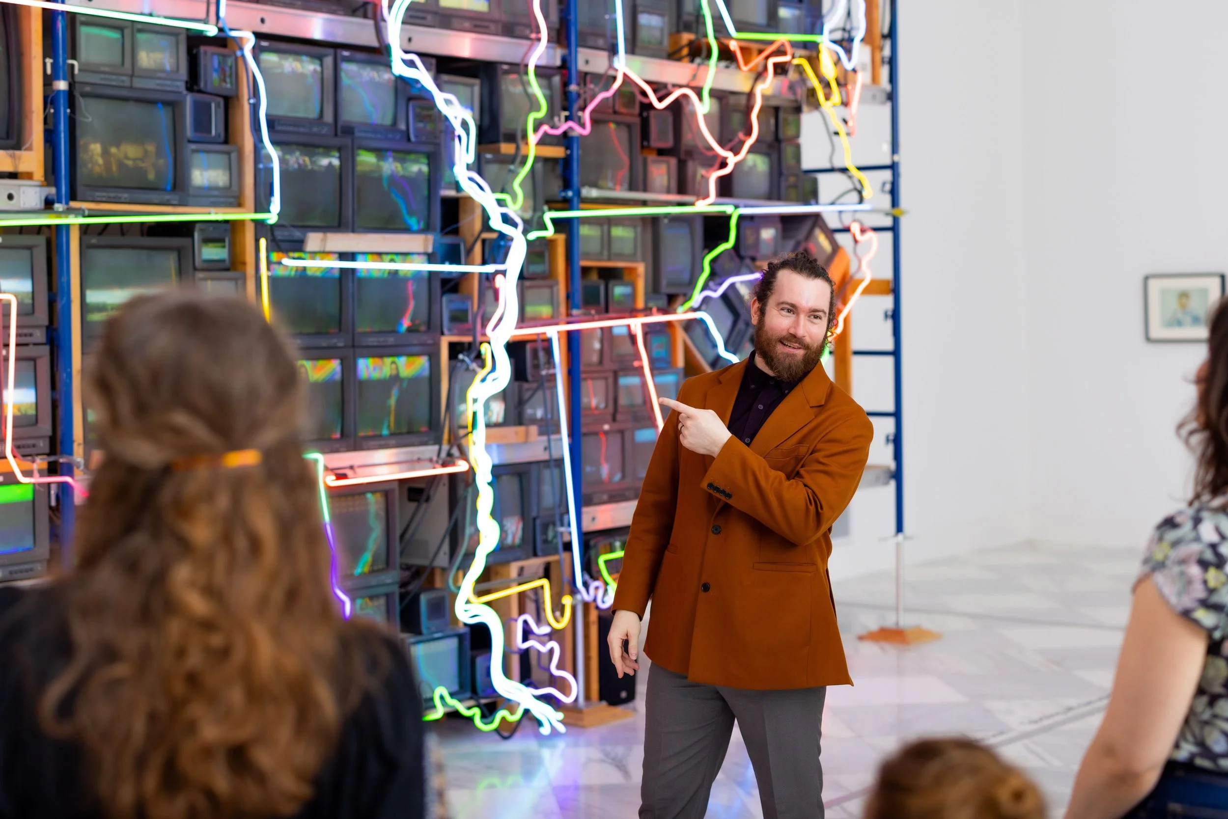 A man with a beard wearing a brown blazer and gray pants is giving a presentation or guided tour to a group of people in front of a colorful neon art installation and vintage televisions.